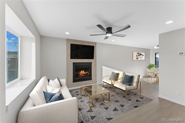 Staged living room with a fireplace, white sofa, geometric rug, and large windows.