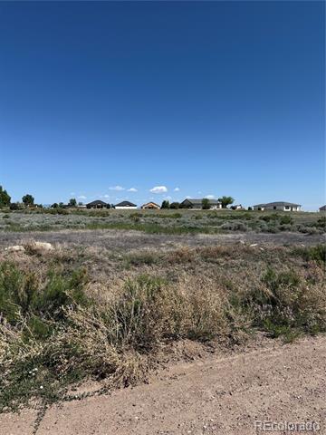 Vacant scrub-covered parcel with a dirt track and distant homes beneath a clear blue sky.