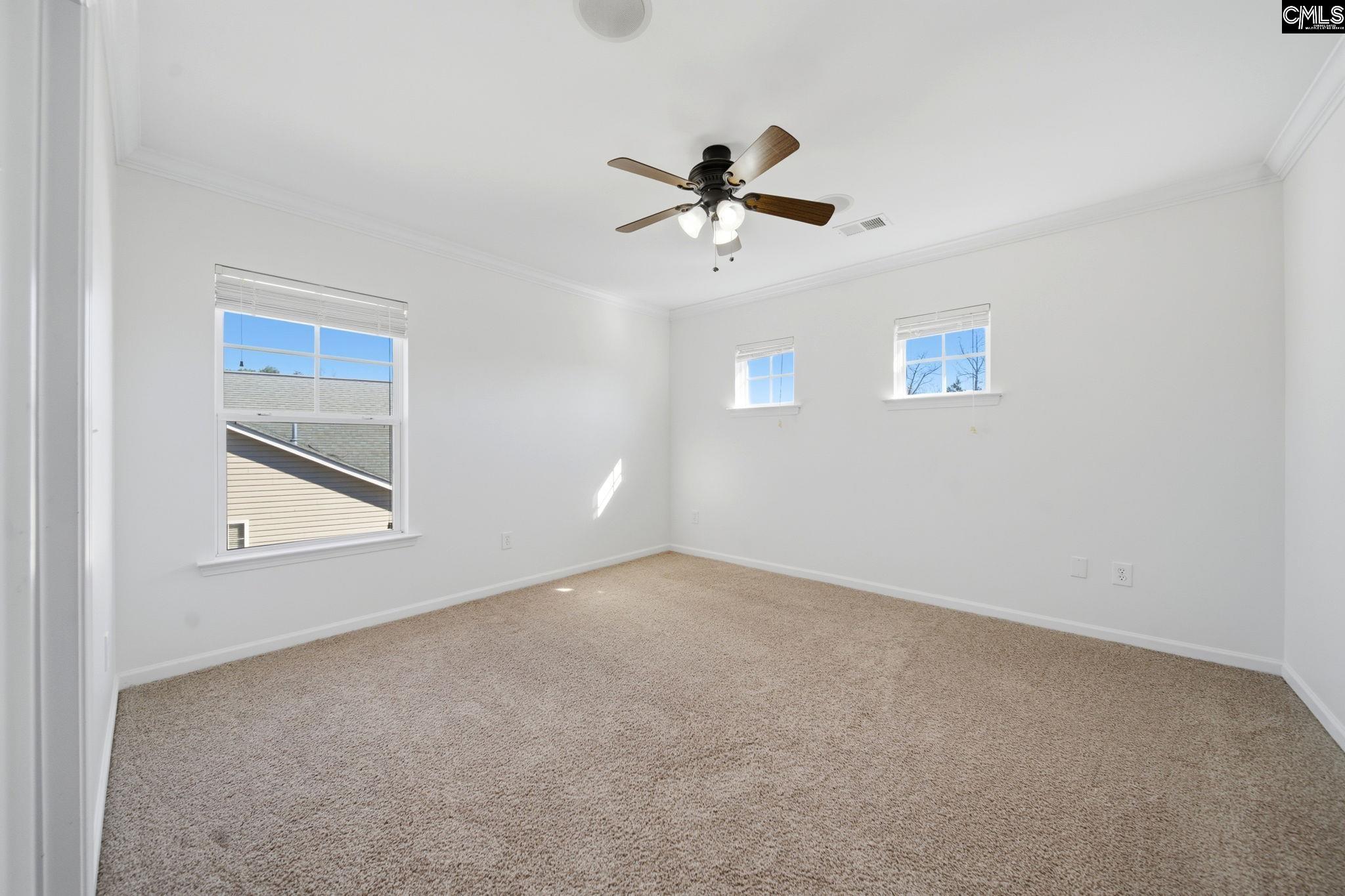 Empty bedroom with beige carpet, three windows, and a ceiling fan with lights.