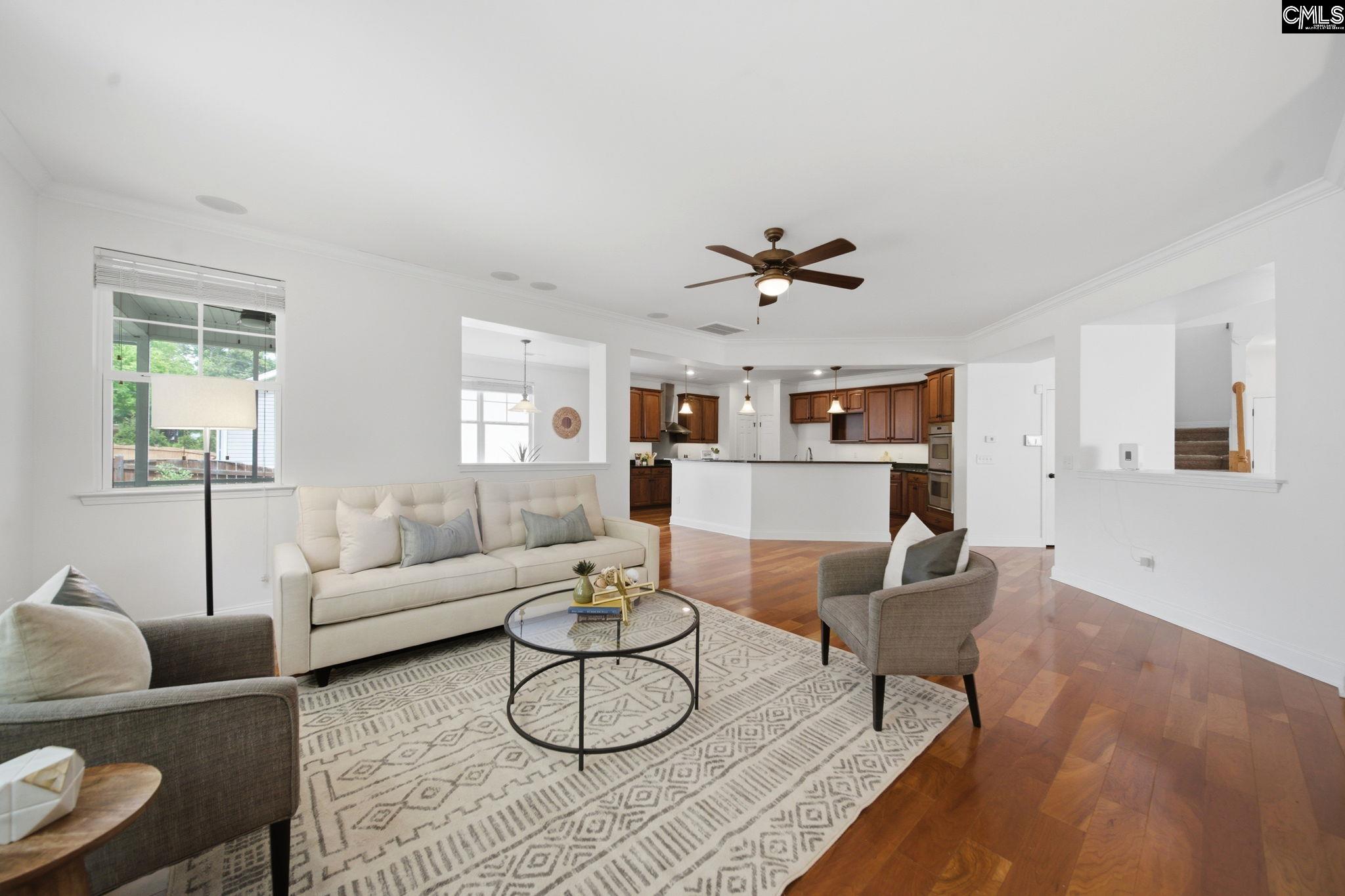 Staged living room with cream sofa, patterned rug, hardwood floors, and an open kitchen with dark wood cabinets.