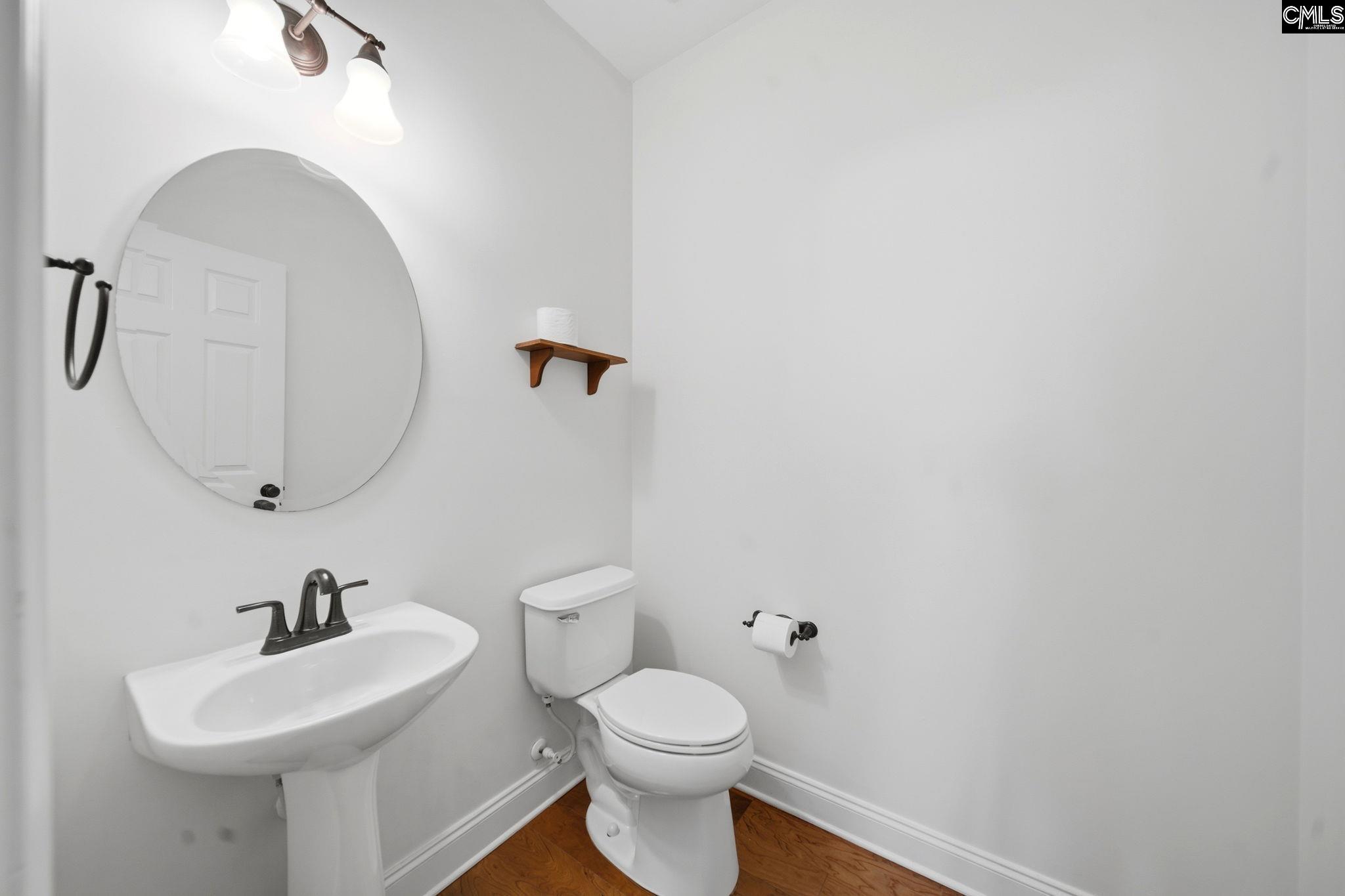 Half bathroom with a pedestal sink, round mirror, toilet, and wood-look flooring.