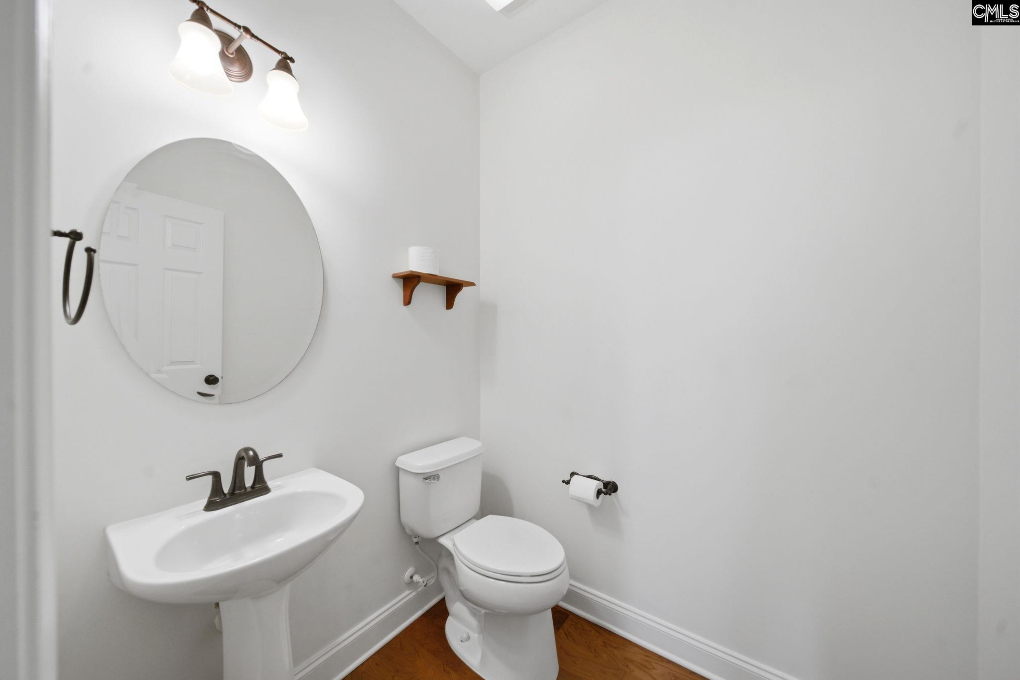 Bathroom with a pedestal sink and round mirror beside a toilet on wood flooring.