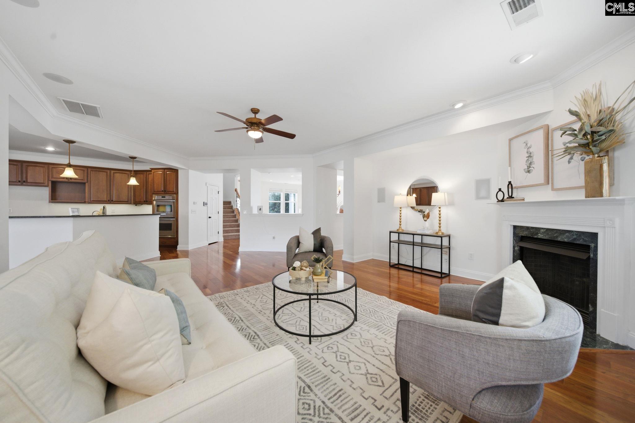 Staged living room with a white fireplace, hardwood floors, and an open kitchen with dark wood cabinets.