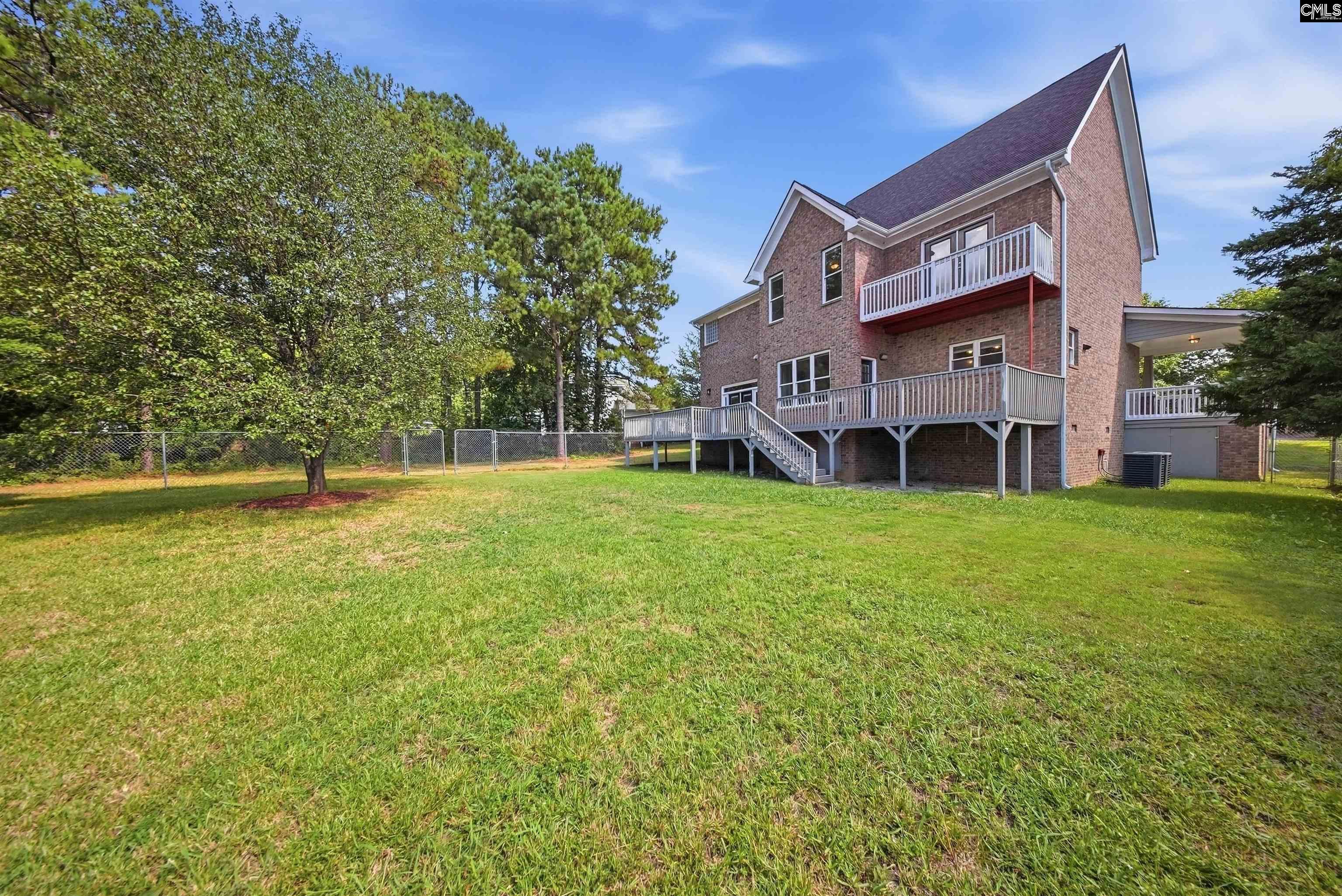 Brick house with elevated decks overlooks a spacious grassy backyard bordered by mature trees and a chain-link fence.