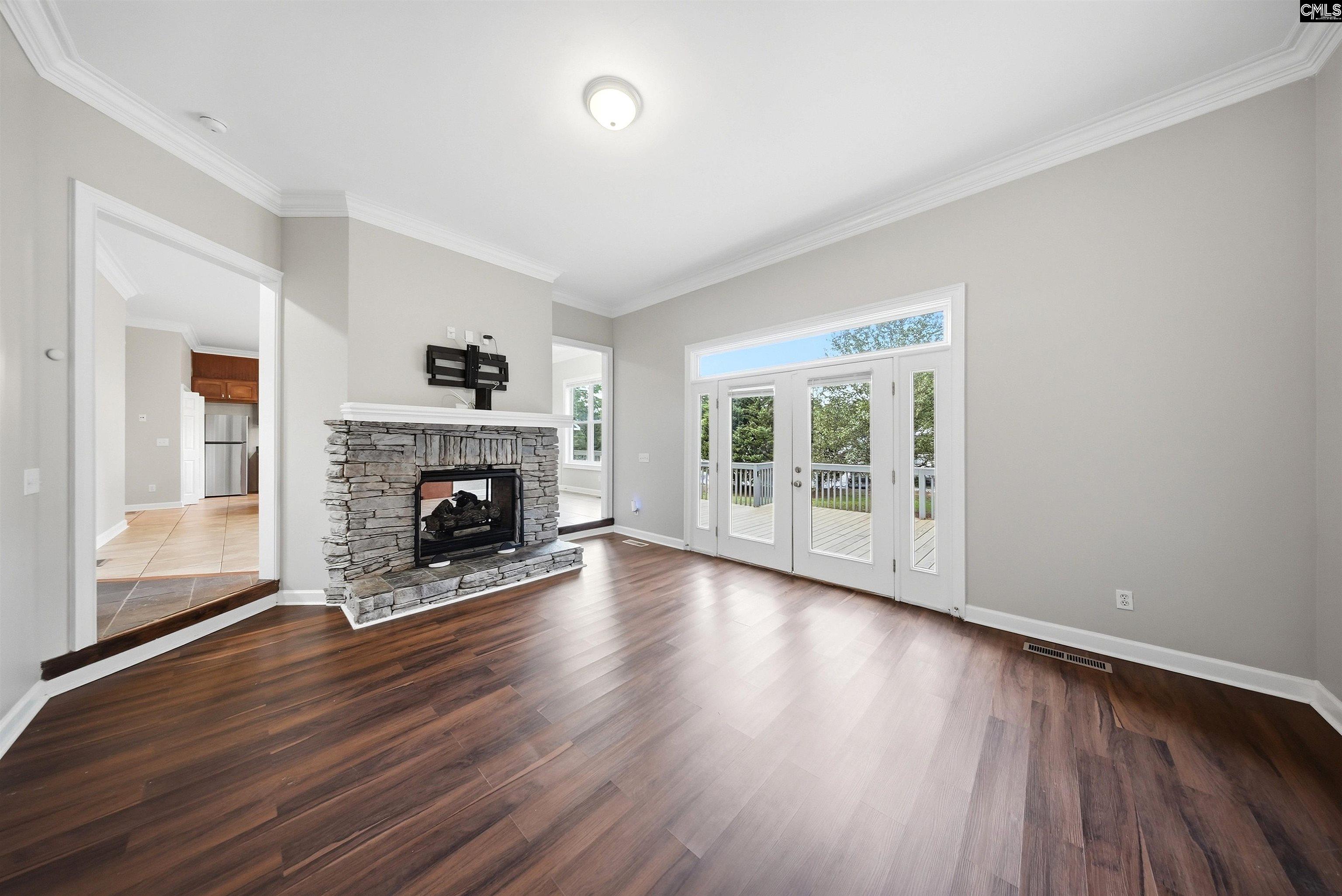 Empty living room with a stone fireplace, dark wood floors, and French doors to a deck.