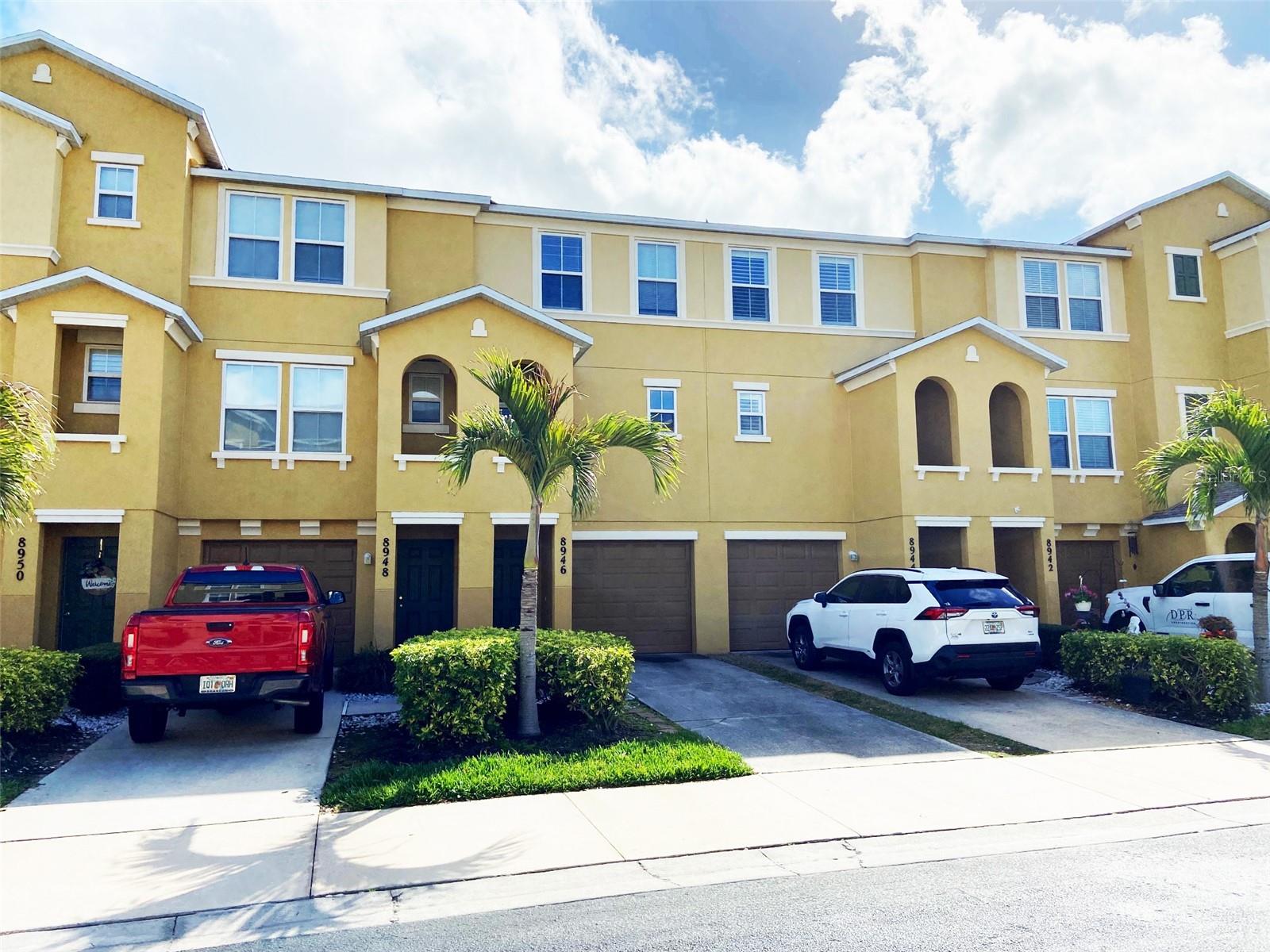 Yellow two-story townhomes with attached garages, palm trees, and parked vehicles along the street.