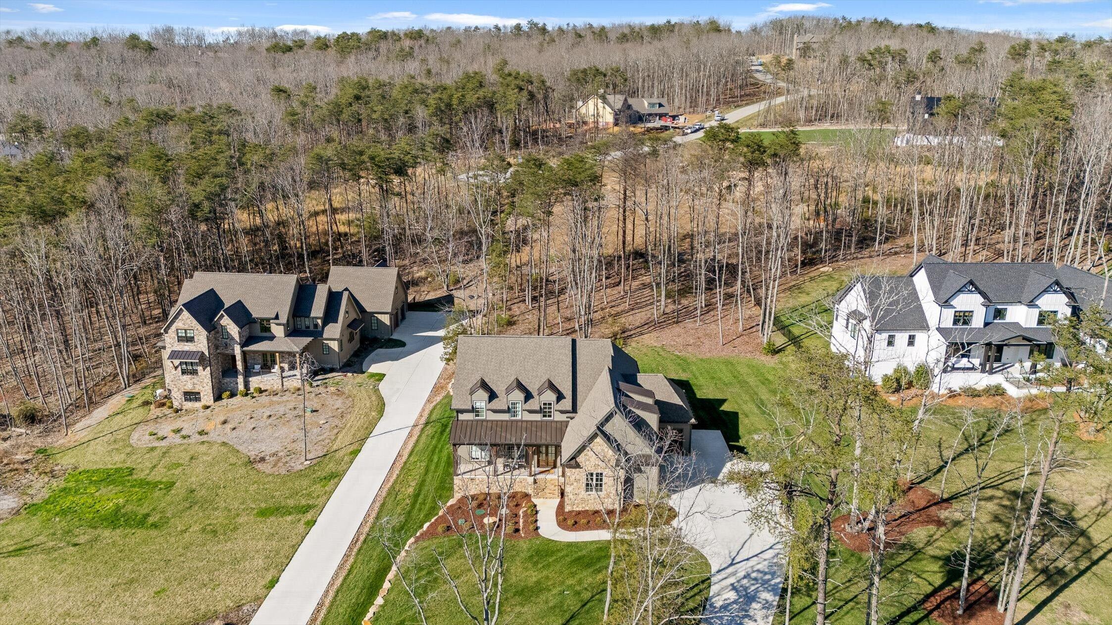 Aerial view of a stone-and-siding home with a curved driveway, lawns, and wooded neighboring properties.