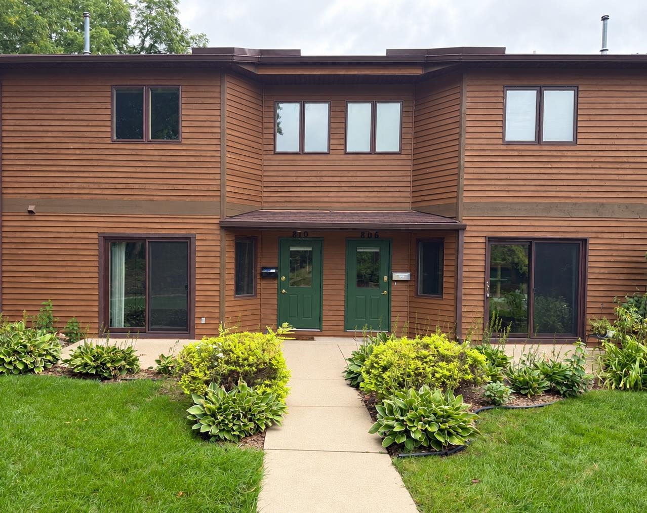 Attached two-story home with green entry doors, wood siding, and a landscaped front walkway.