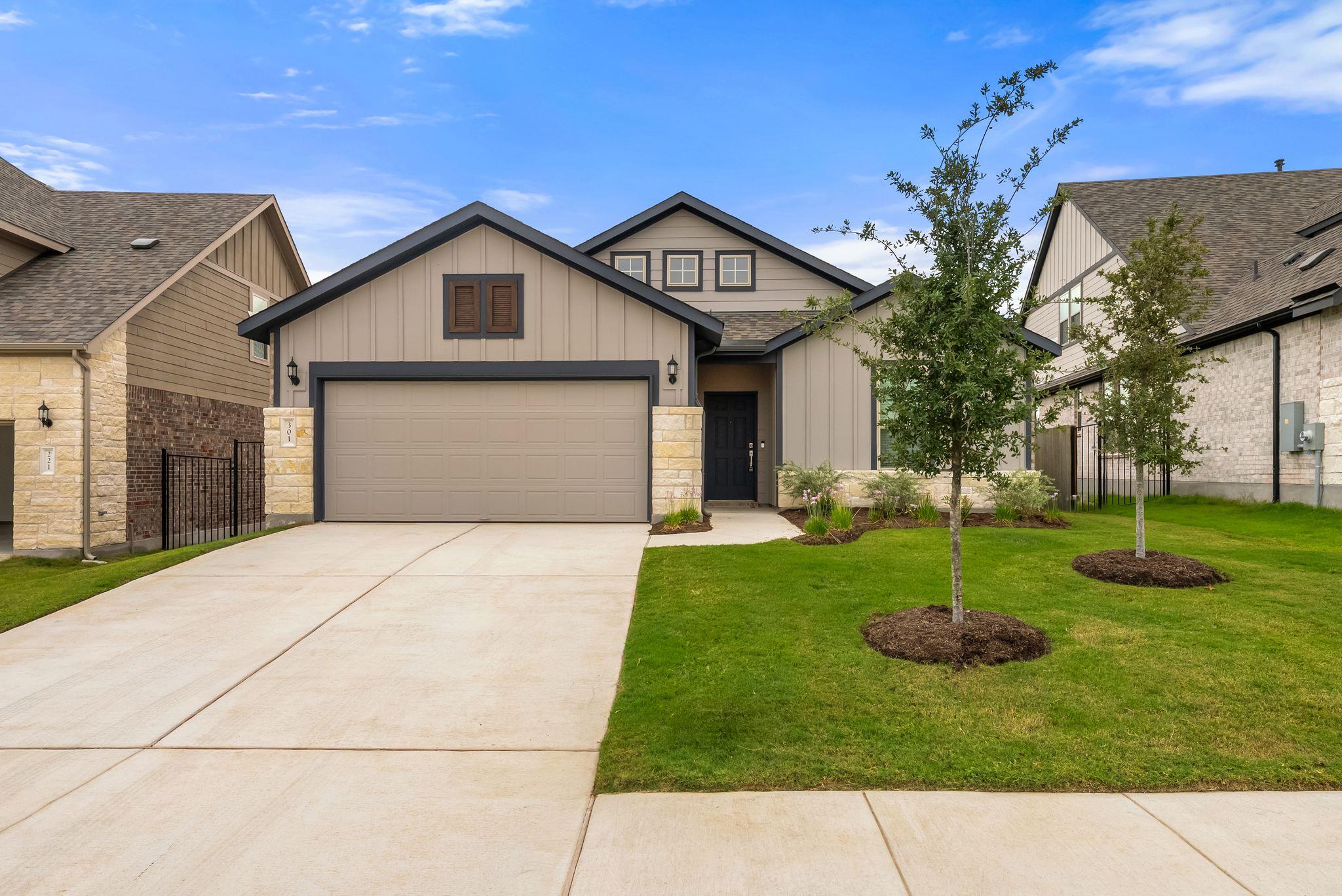 Beige gabled home with a two-car garage, wide driveway, and young trees on the lawn.
