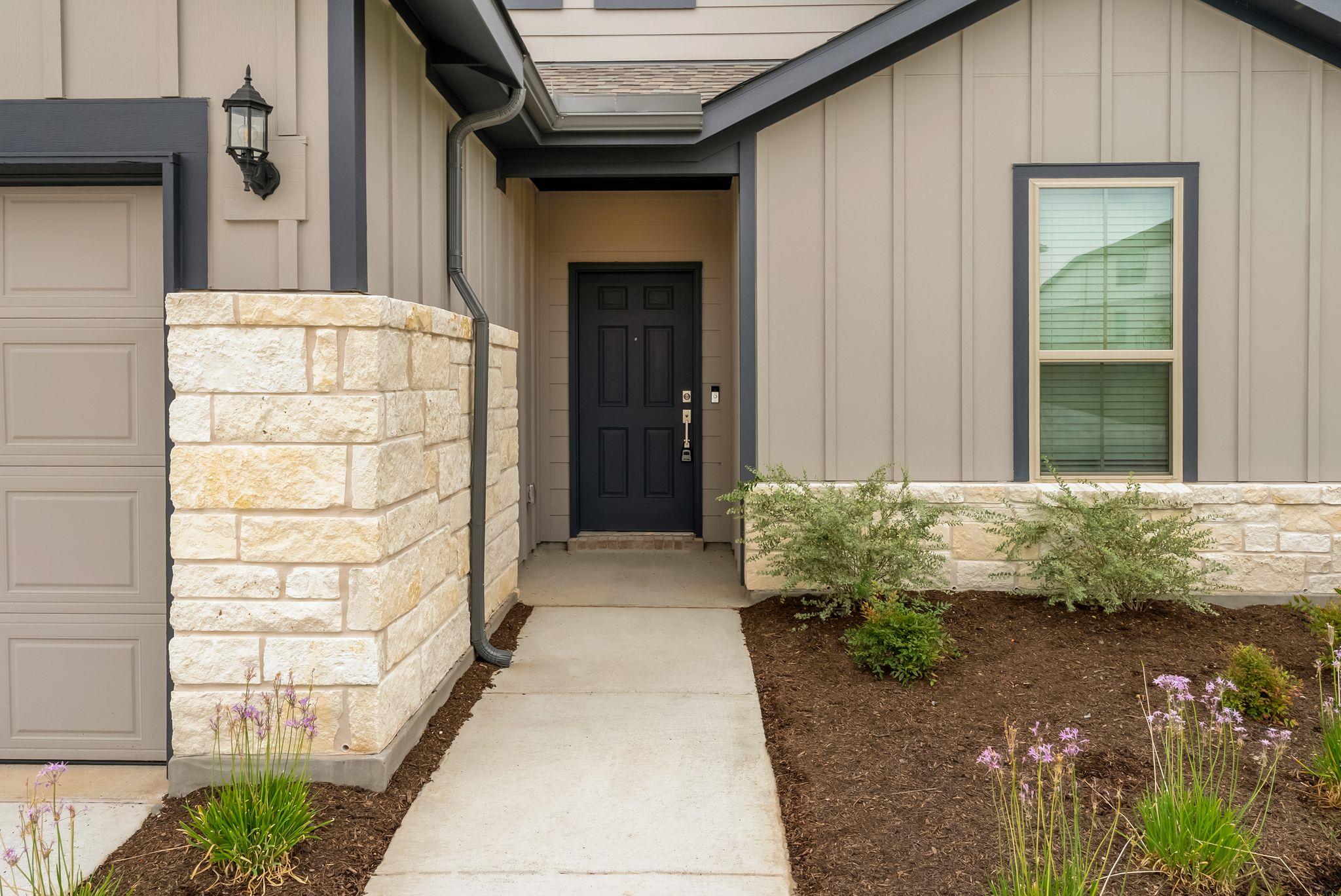 Covered home entry with a dark front door, pale stone walls, and flower beds along the walkway.
