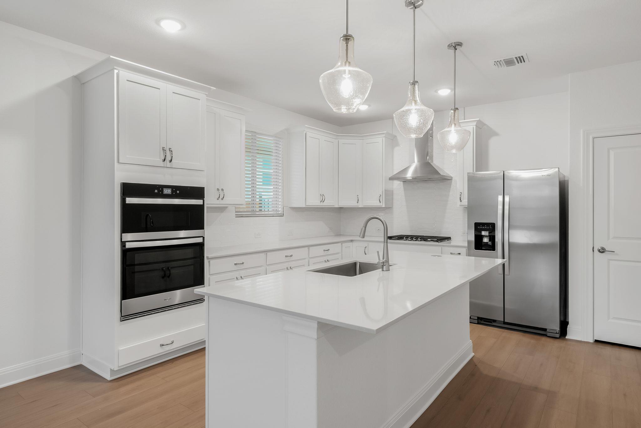 Bright white kitchen with a large island, double ovens, stainless steel appliances, and glass pendant lights.