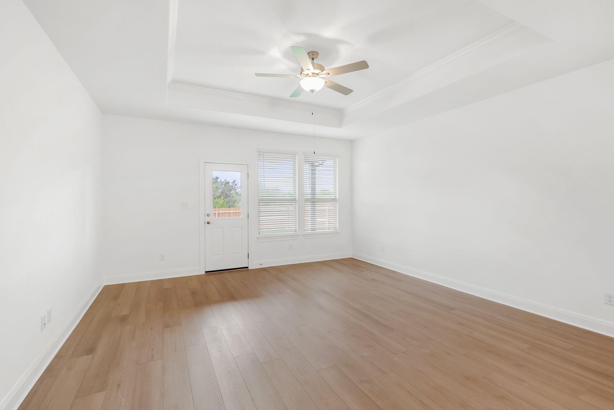 Empty room with light wood flooring, a tray ceiling, ceiling fan, and windows beside a glass-paneled door.