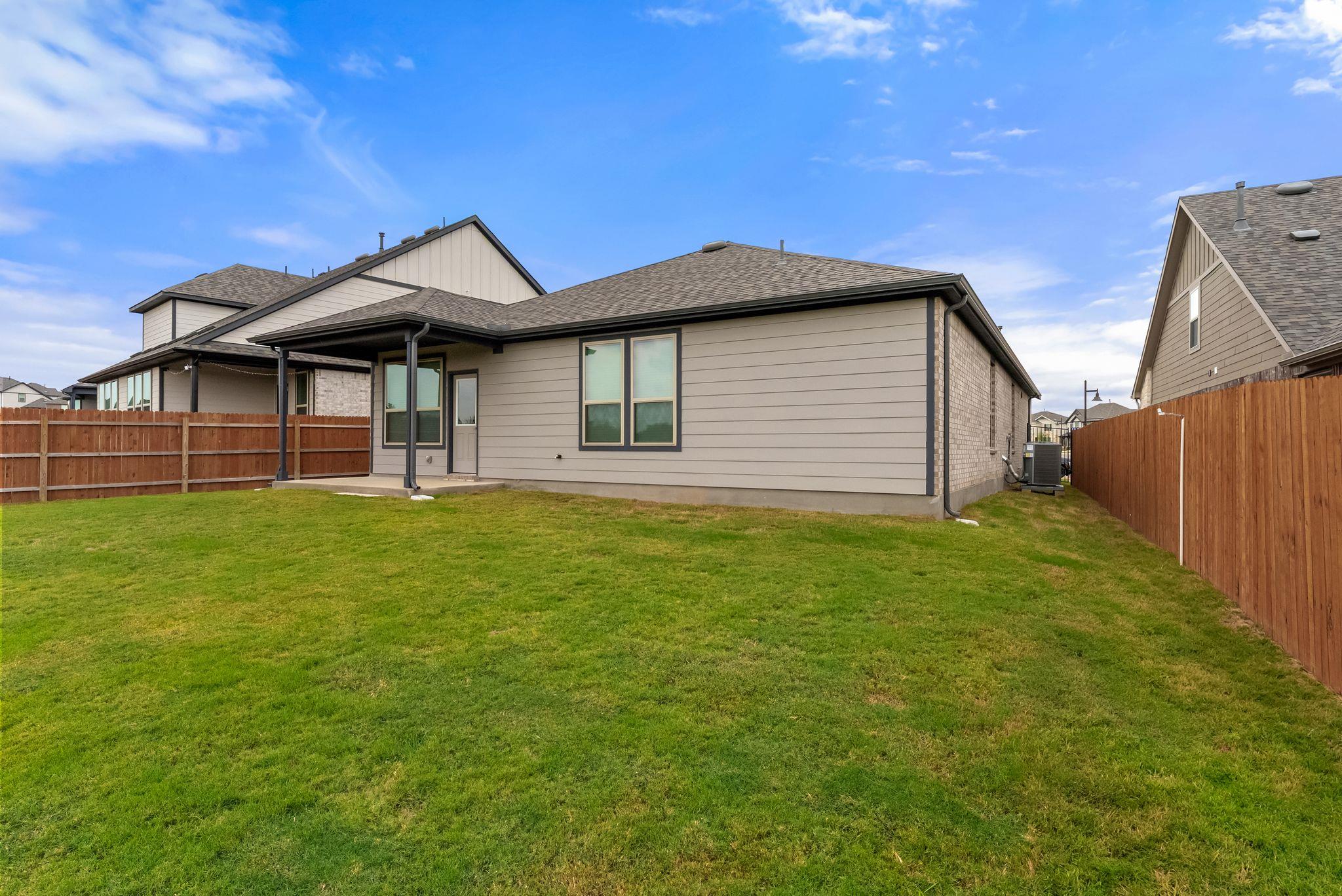 Rear of a gray-sided house with a covered patio, fenced backyard, and broad green lawn.