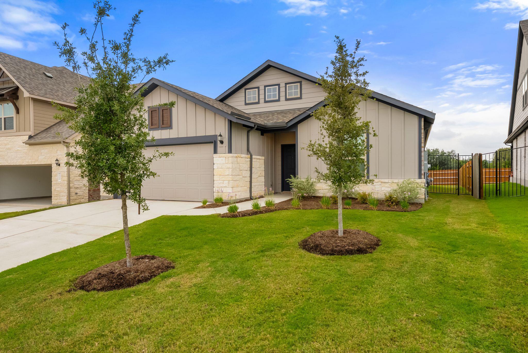 Beige home with a two-car garage, stone accents, young trees, and a fenced side yard.