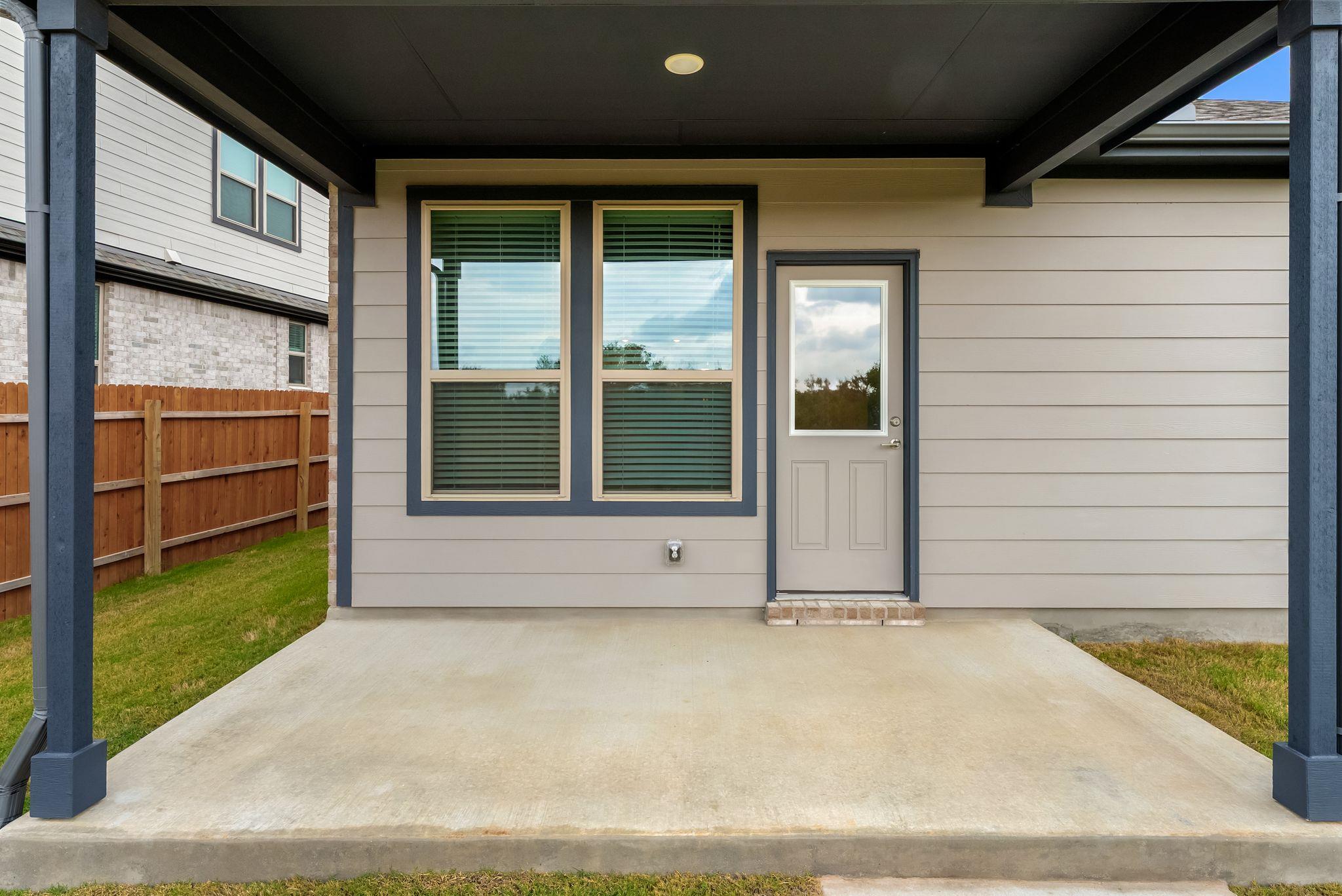 Covered concrete patio with a gray door, two large windows, and dark support posts.