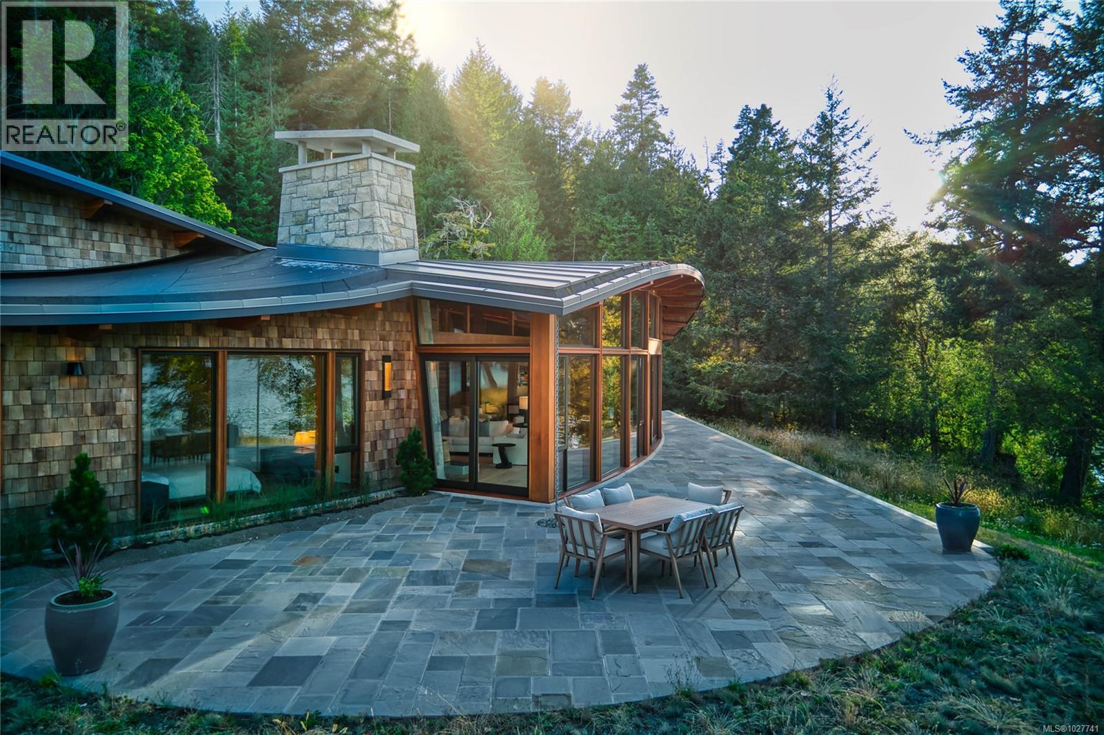 Modern wood-shingle home with a broad stone patio and outdoor dining table among evergreen trees.