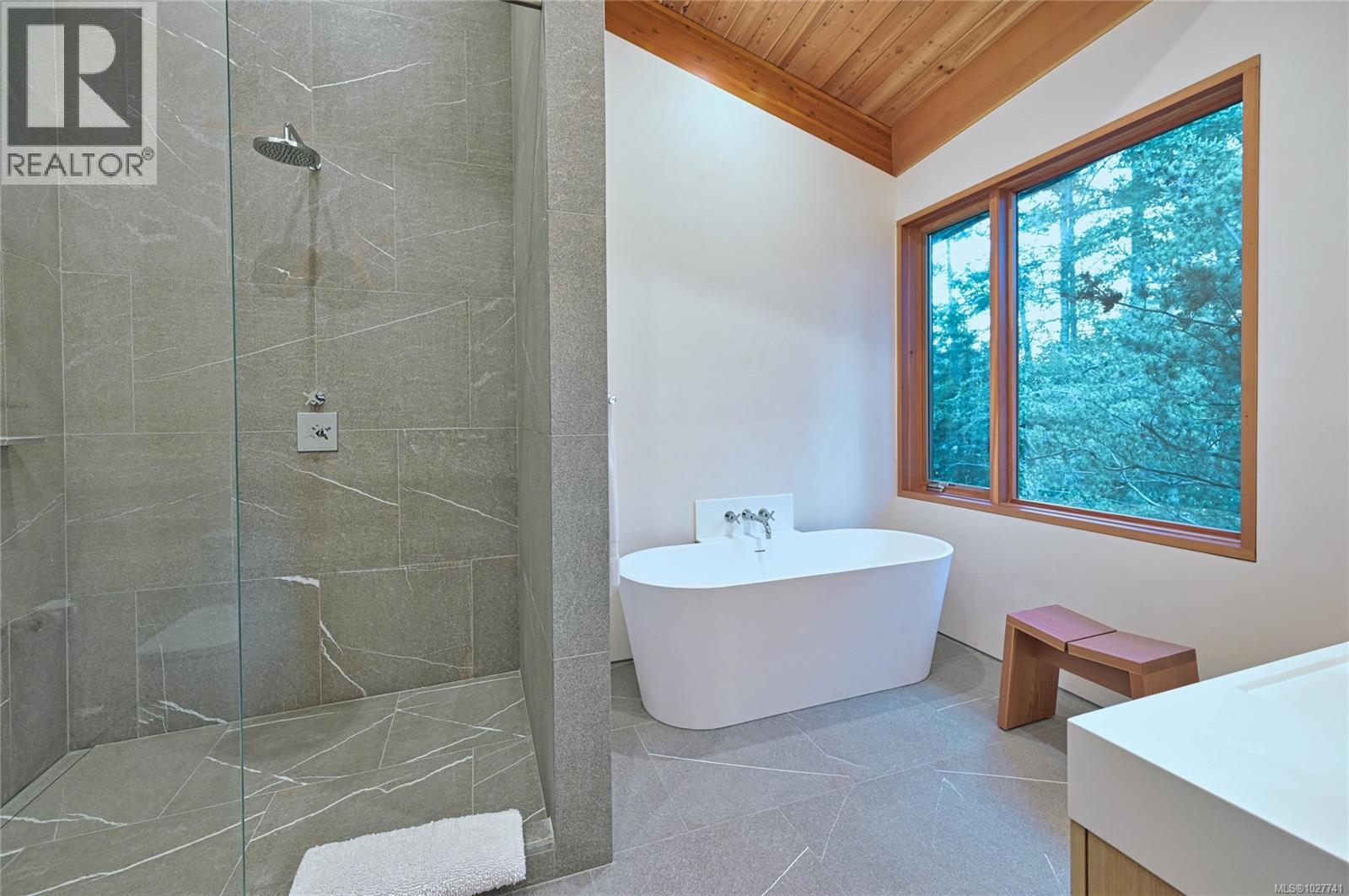 Bathroom with a stone shower, freestanding tub, and wood-framed window overlooking trees.