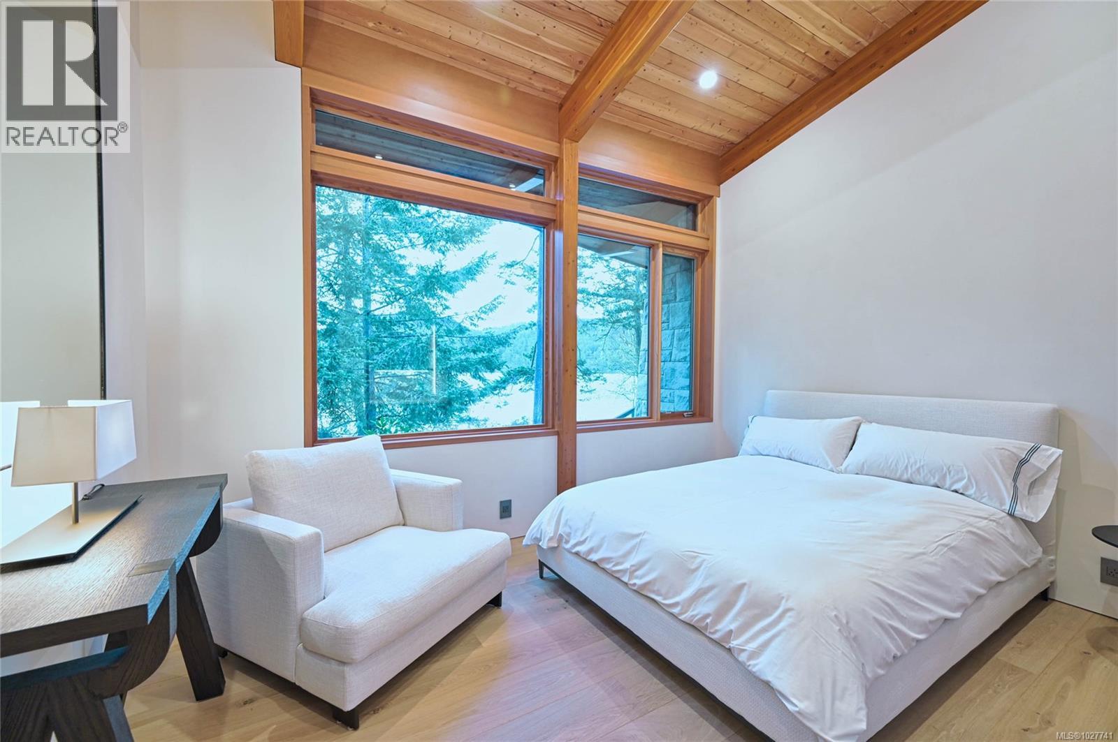 Bedroom with a white bed, exposed wood ceiling, and large windows overlooking trees and water.