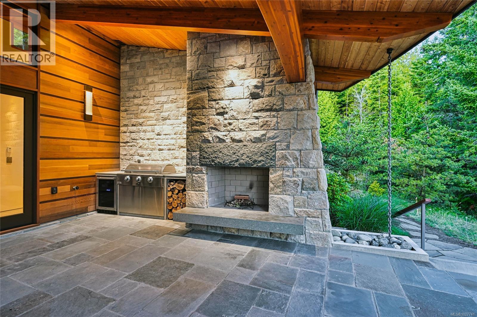 Covered patio with a massive stone fireplace, built-in grill, and forest views.