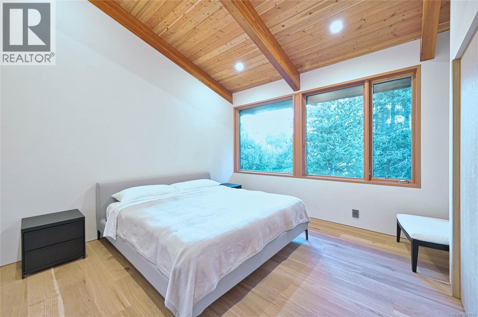 Bedroom with a white bed, vaulted wood ceiling, and large windows overlooking trees.
