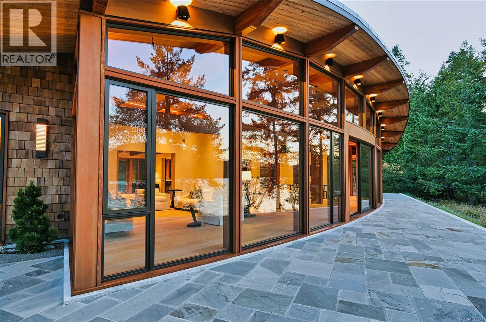 Modern home with curved timber roof, expansive windows, and a stone patio beside trees.