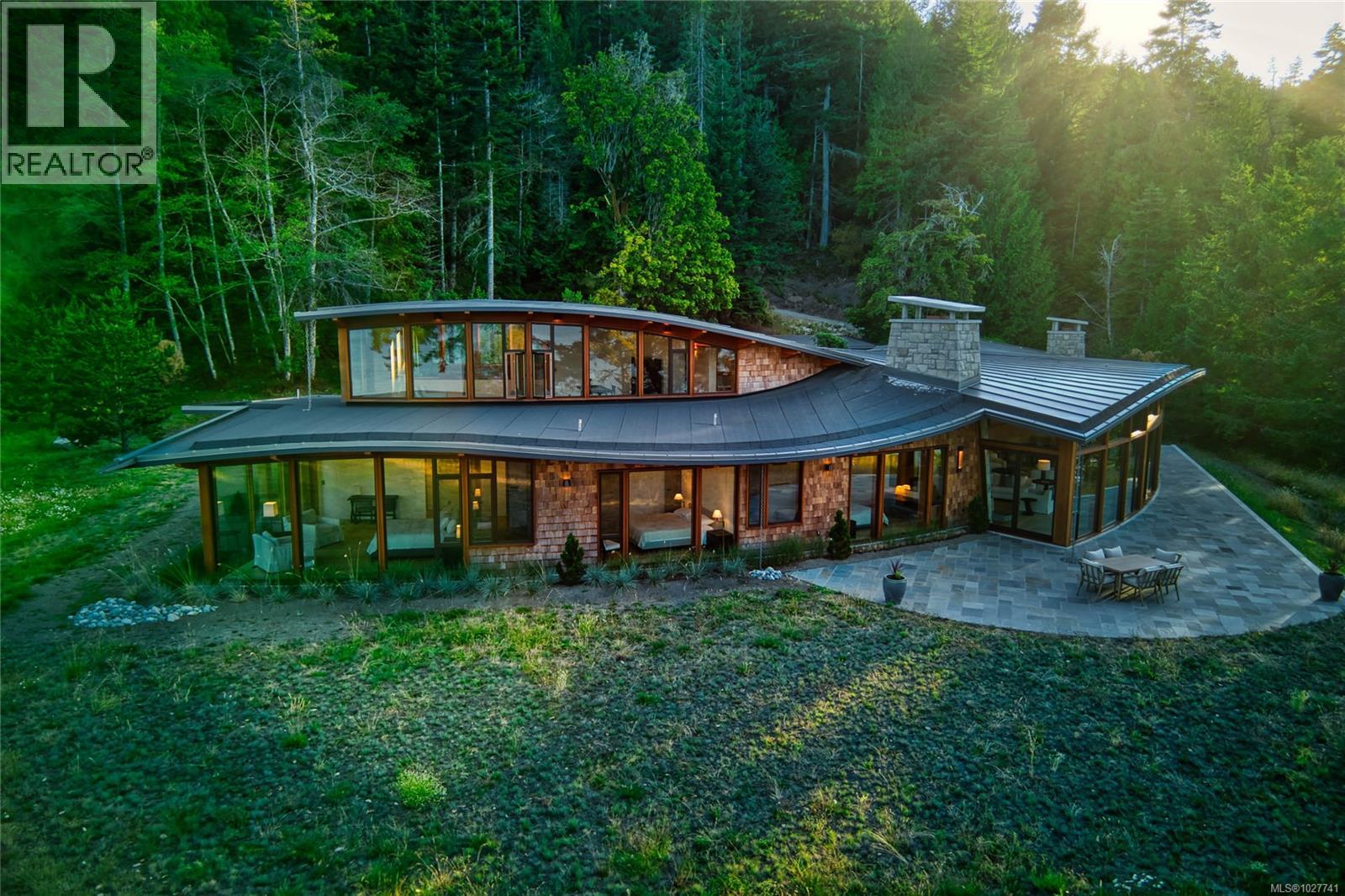 Modern curved-roof home with expansive windows and a stone patio in a forest clearing.