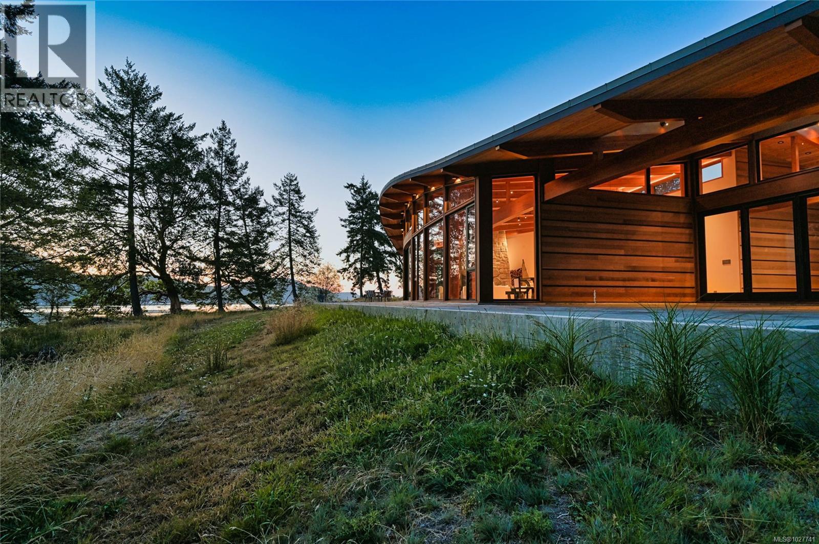 Modern wood-clad home with curved roof and pool beside a tree-lined waterfront at dusk.
