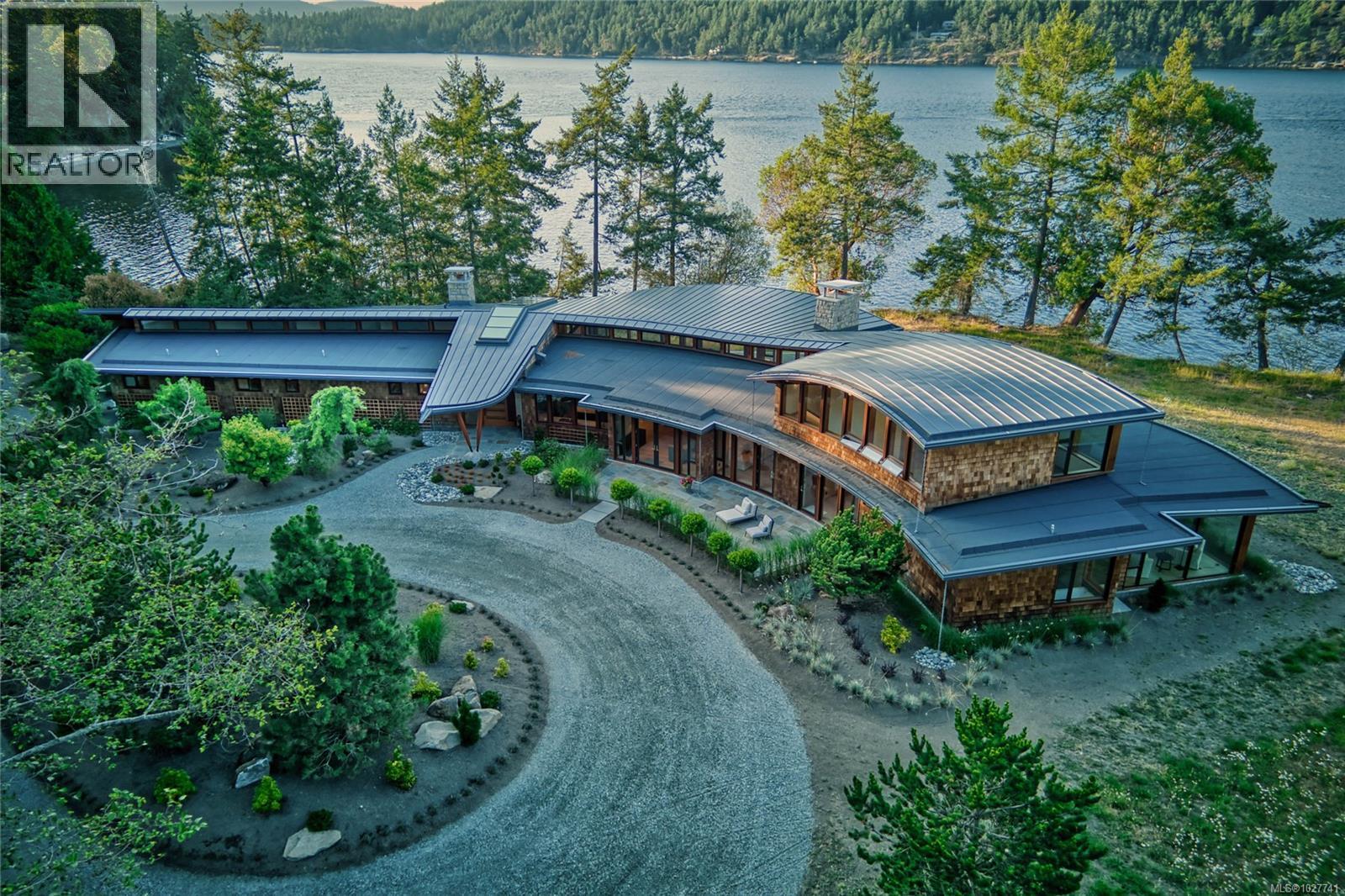 Aerial view of a modern waterfront home with sweeping metal roofs and a curved gravel driveway beside a forested bay.