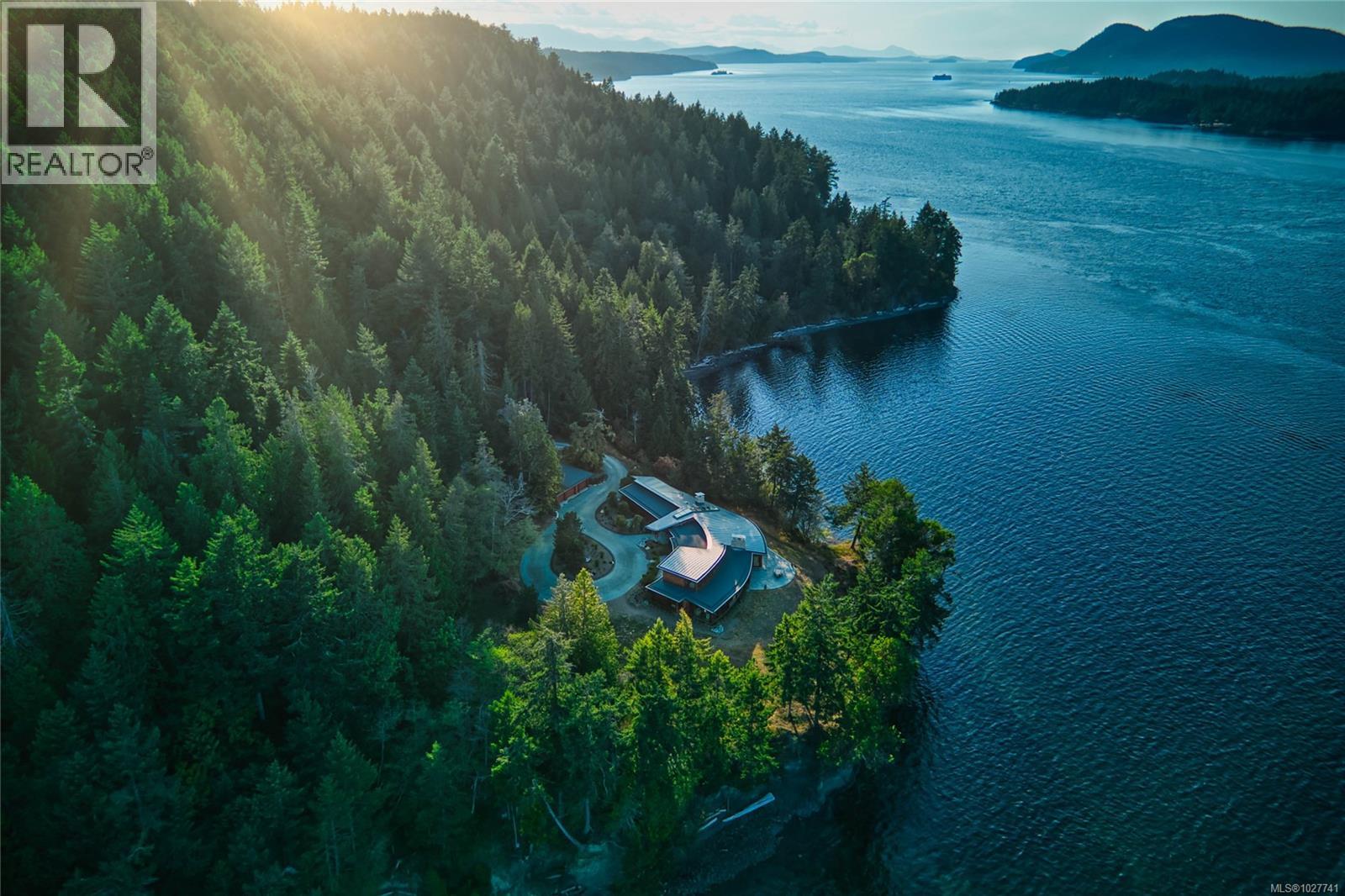 Aerial view of a modern waterfront home surrounded by forest beside a blue inlet.