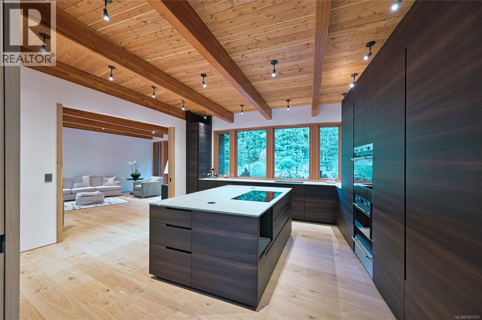 Modern kitchen with a large island, dark cabinetry, exposed wood ceiling, and windows overlooking trees.
