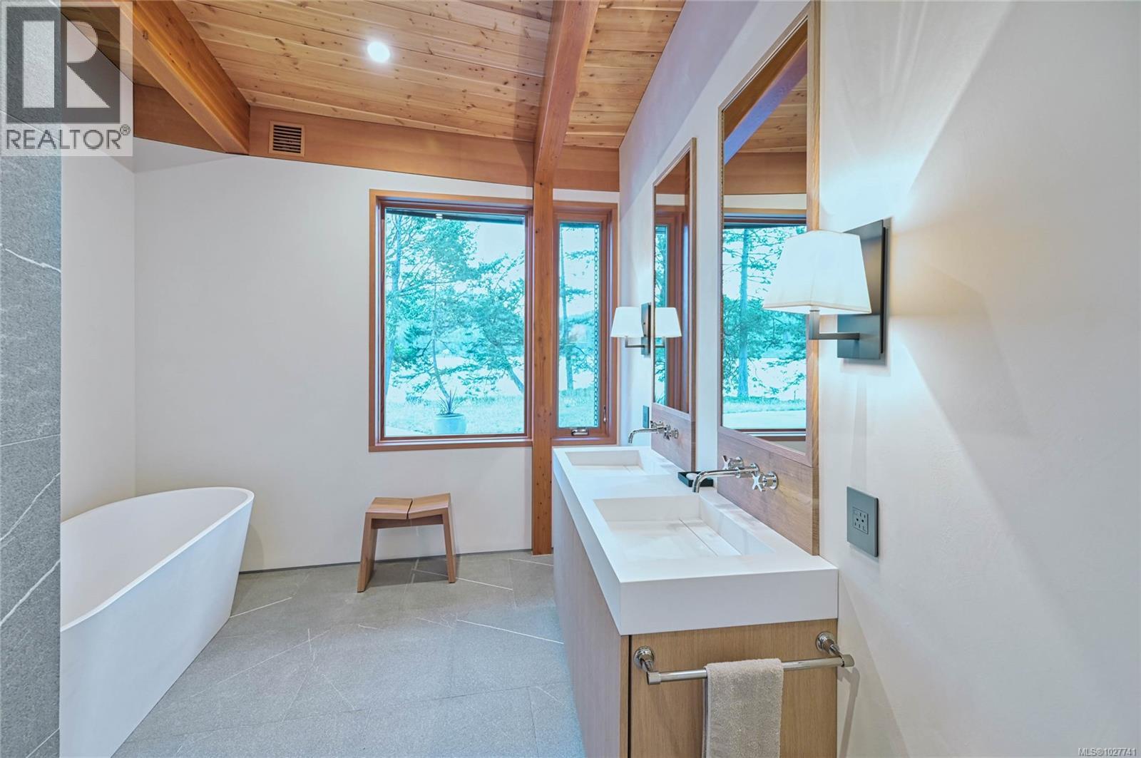 Bathroom with a freestanding tub, double vanity, vaulted wood ceiling, and windows overlooking water.