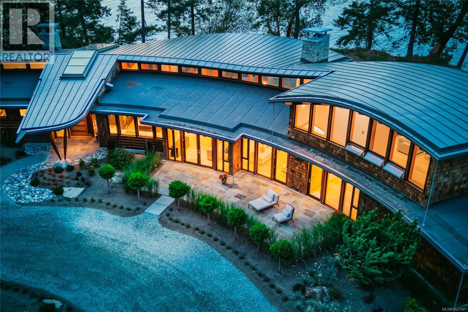 Modern waterfront home with sweeping metal roofs and glowing windows beside a stone patio.