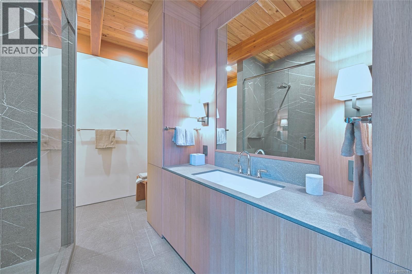 Modern bathroom with a glass shower, gray stone tile, and a wide wood-fronted vanity beneath a vaulted wood ceiling.