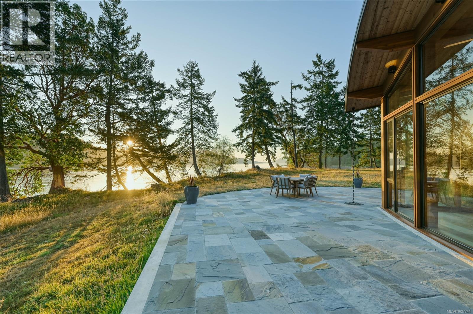 Waterfront stone patio beside a modern home, with outdoor dining and sunset through evergreen trees.