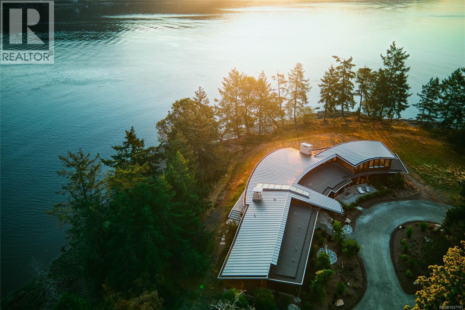 Aerial view of a curved-roof waterfront home among trees beside calm water.