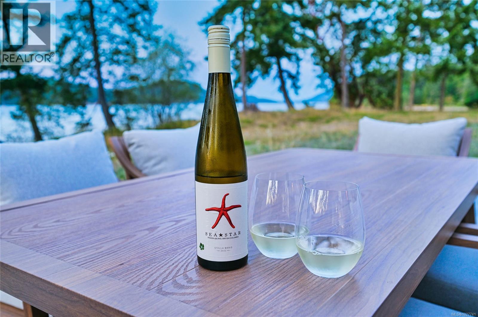 Wine bottle and two glasses on a patio table beside cushioned chairs, trees, and waterfront.
