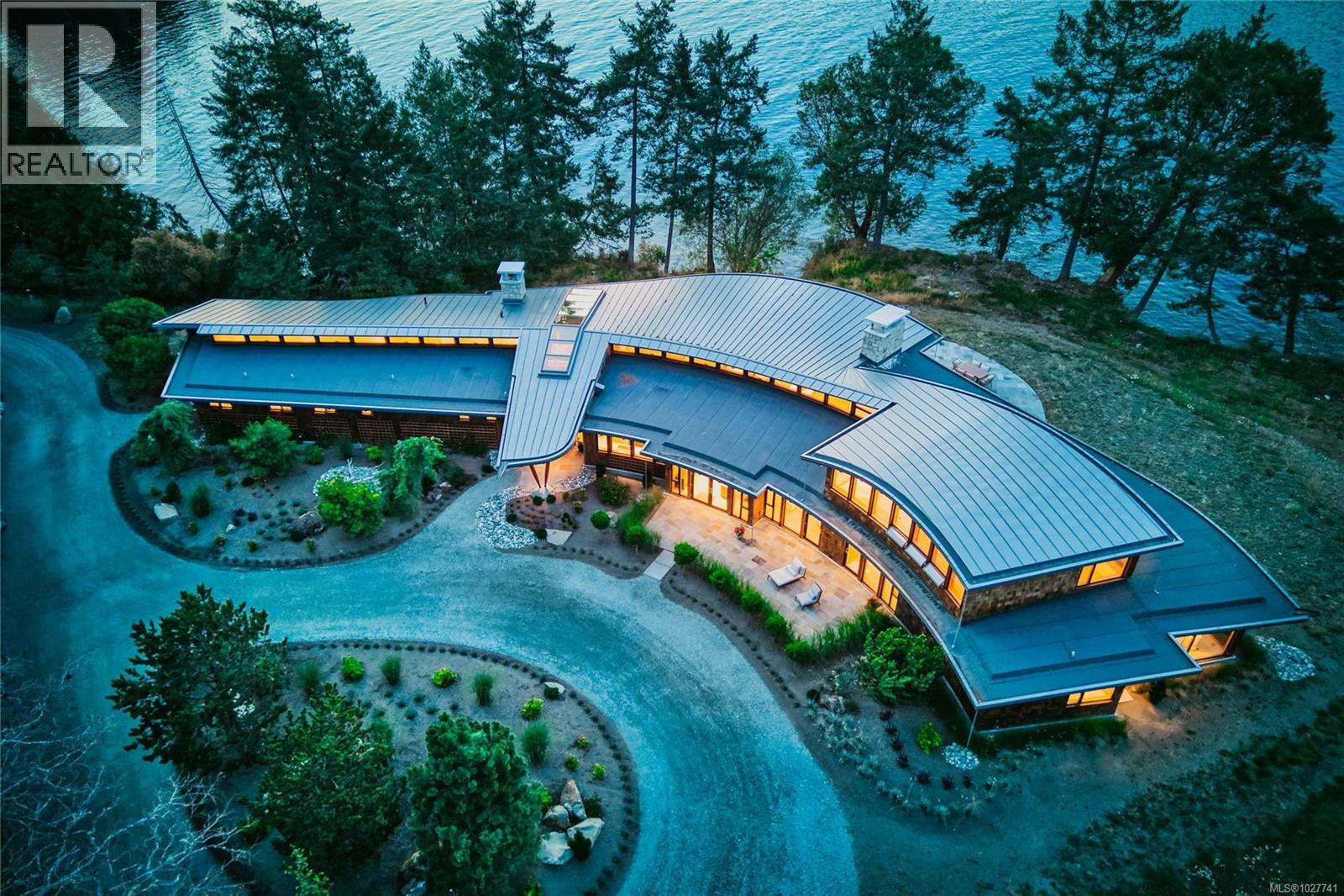 Aerial view of a modern waterfront home with curved metal roofs and warmly lit windows.
