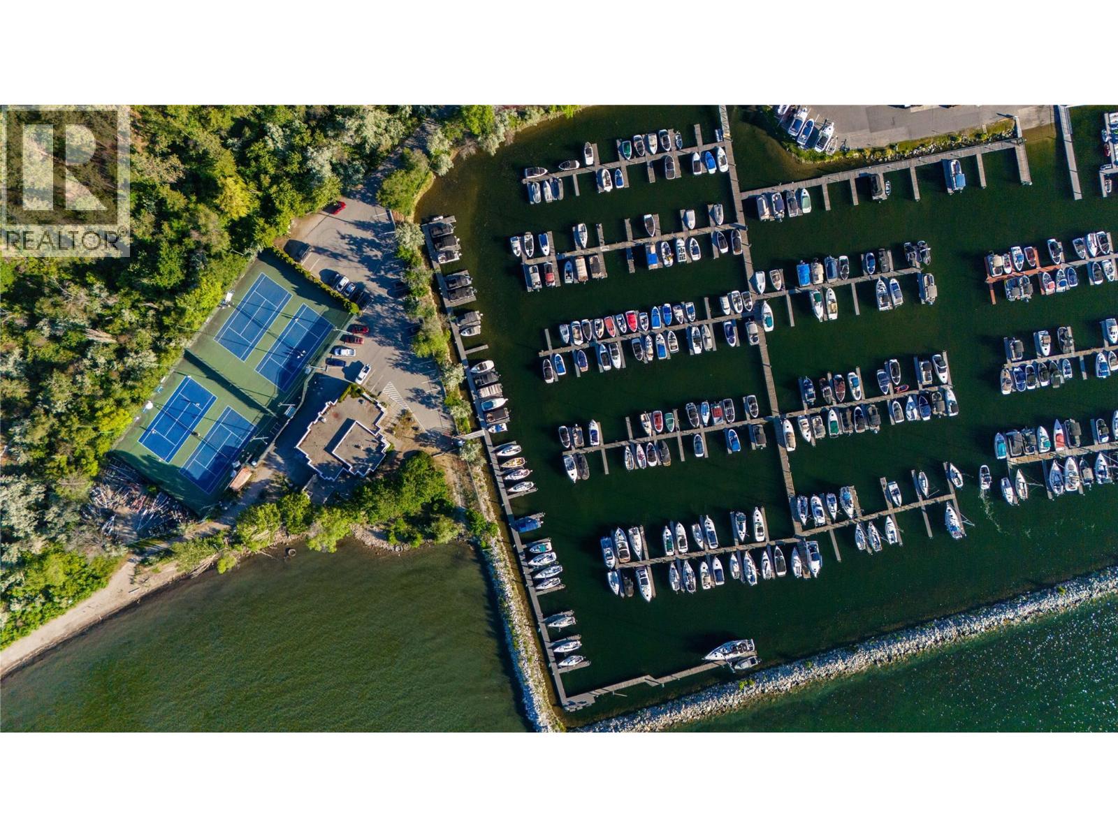 Aerial view of a marina with rows of boats, two tennis courts, and a tree-lined shoreline.