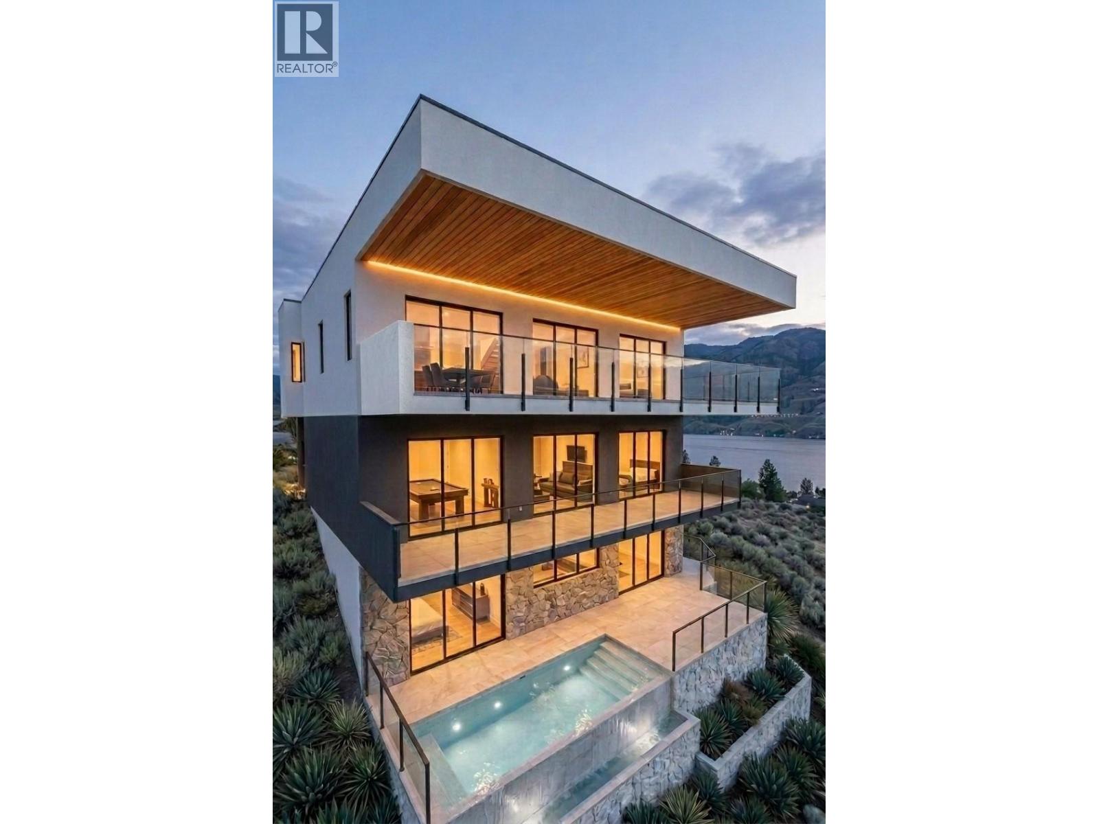 Modern multi-level home with glass terraces and a pool overlooking water and mountains at dusk.