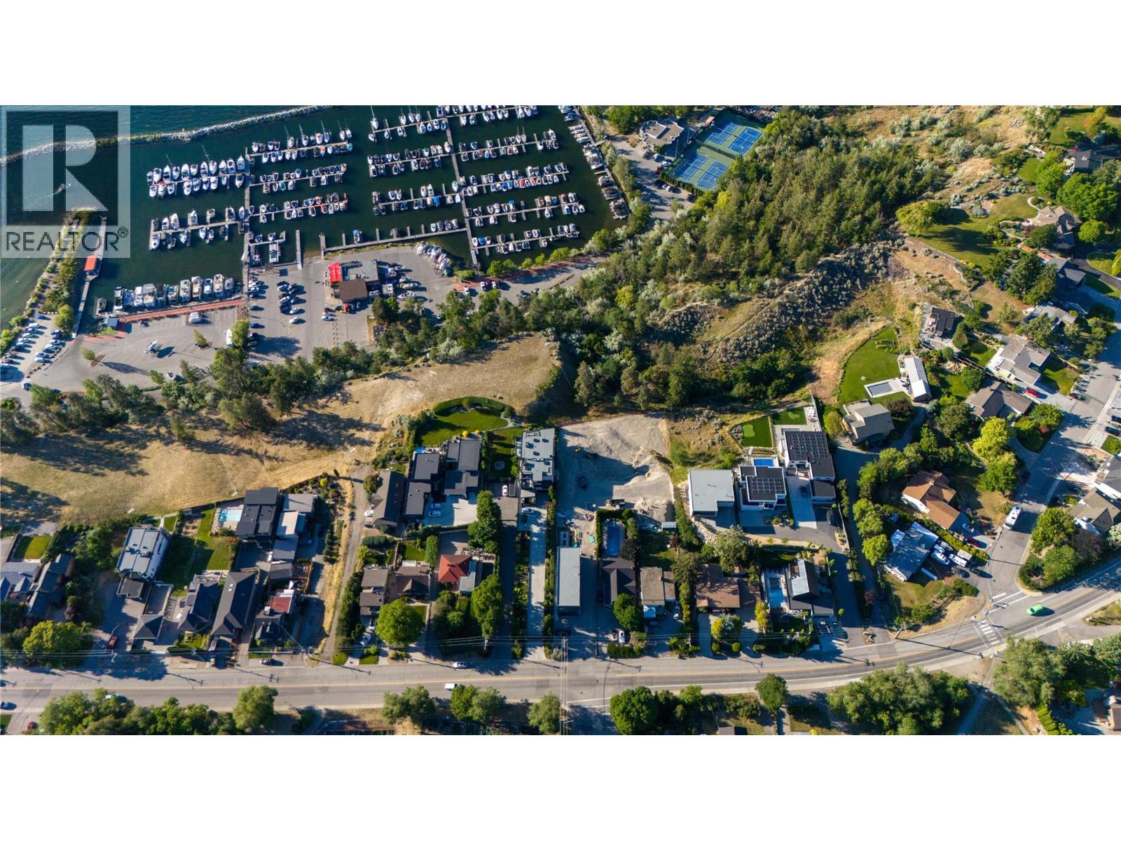 Aerial view of homes beside a marina filled with boats, with tennis courts and a wooded hillside nearby.