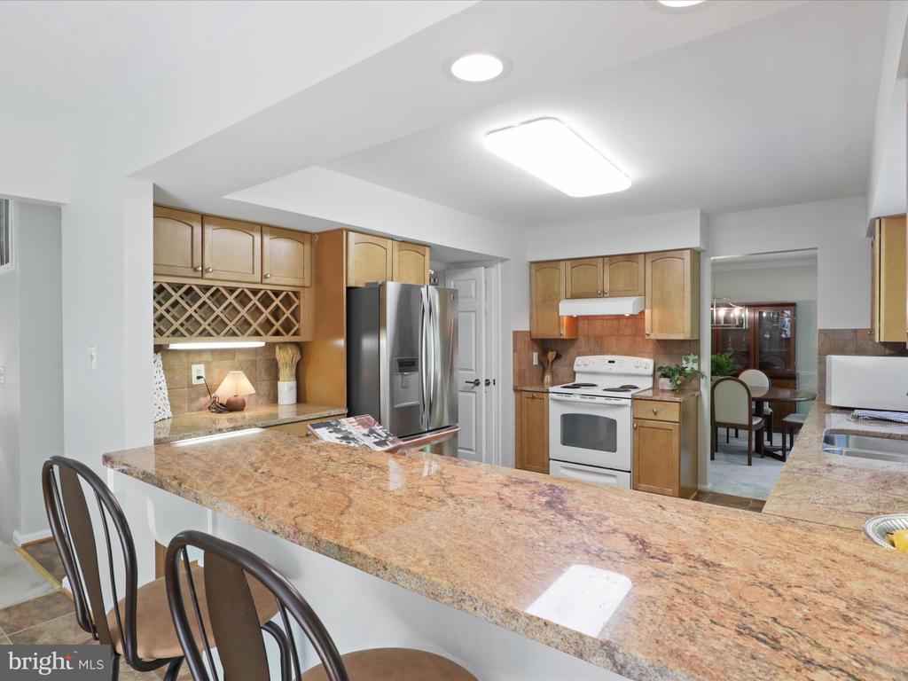 Kitchen with wood cabinets, stainless steel refrigerator, and a stone breakfast bar with dark stools.