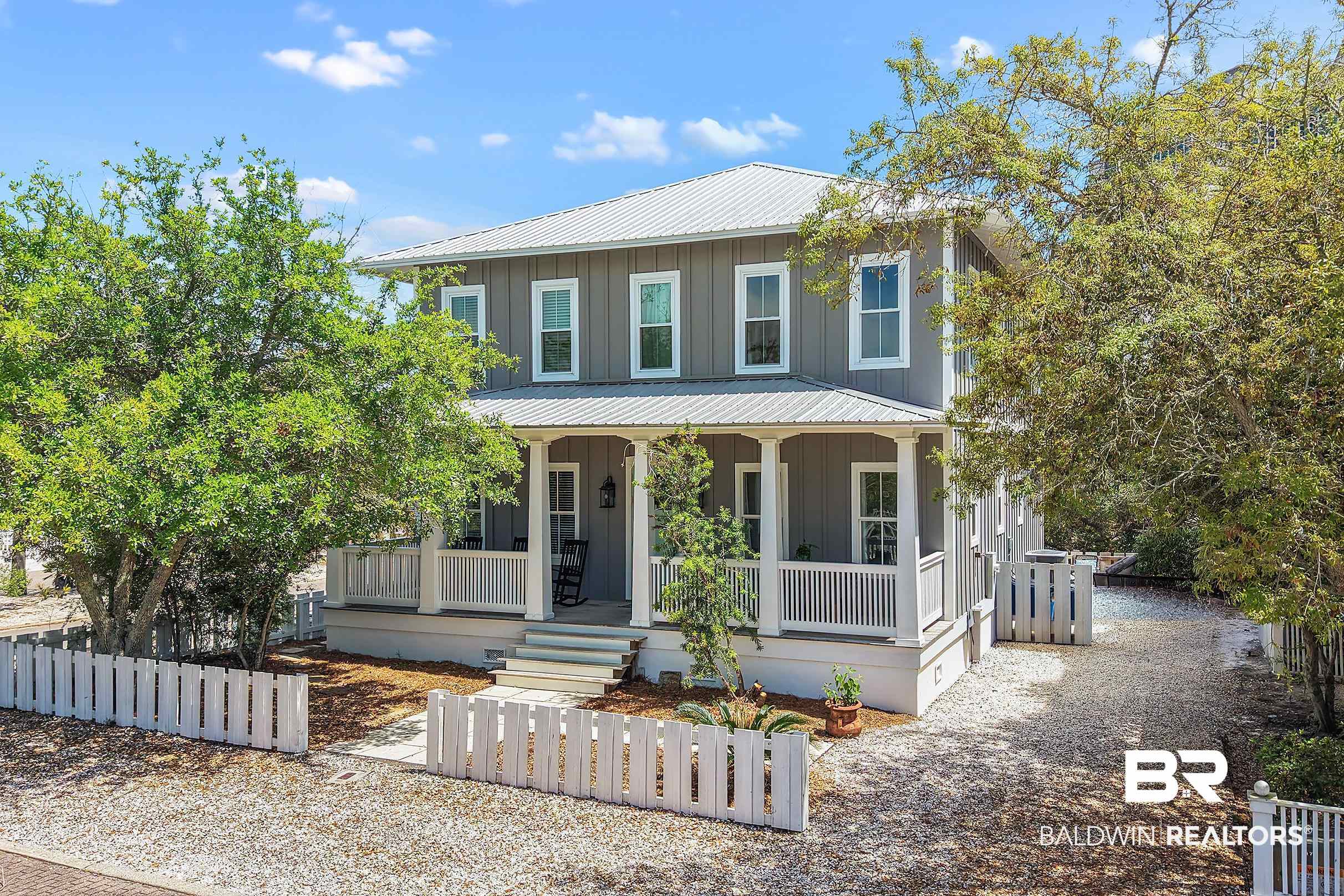 Two-story gray house with a covered porch, white picket fence, and gravel driveway.