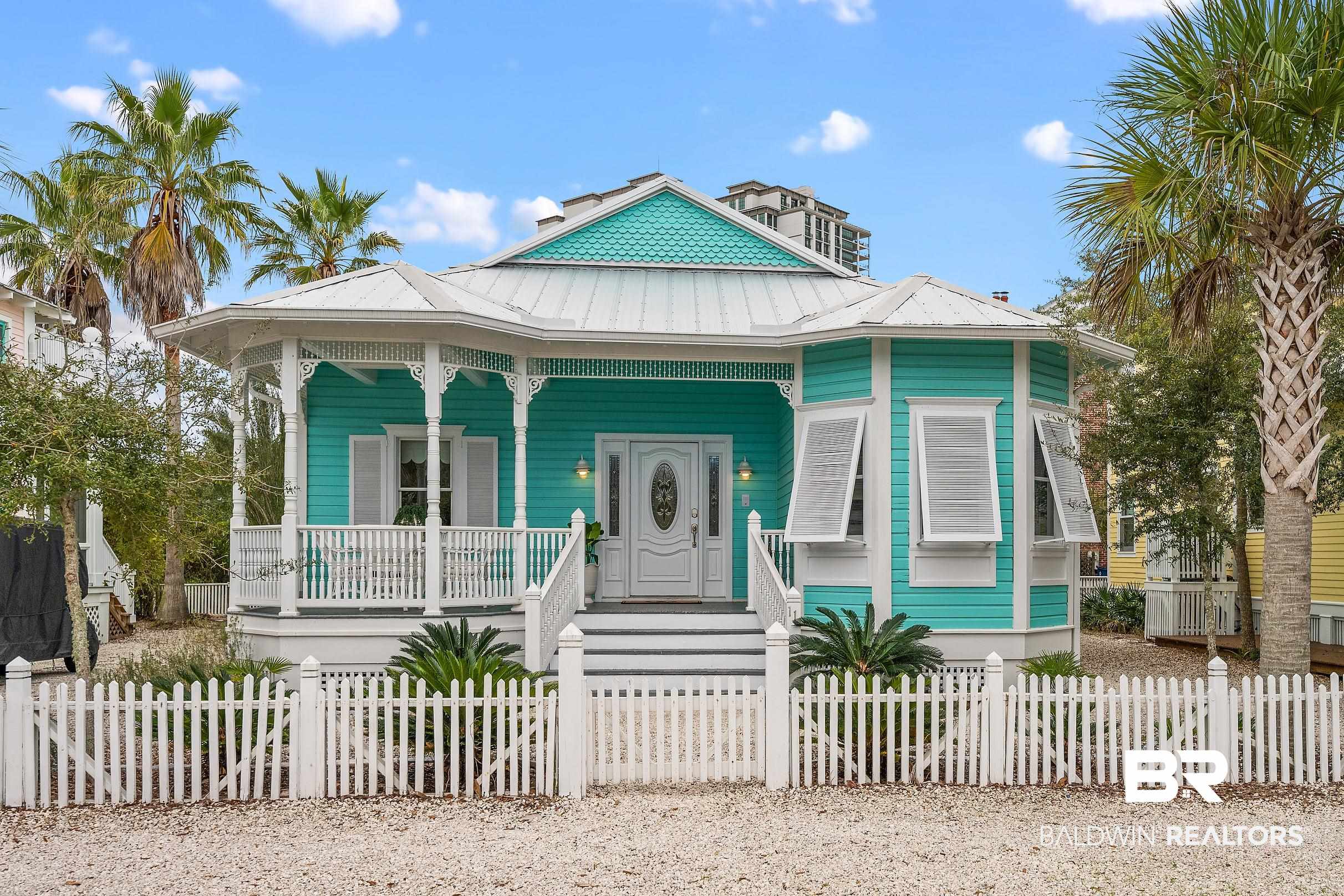 Turquoise coastal cottage with a white metal roof, ornate porch, and white picket fence.