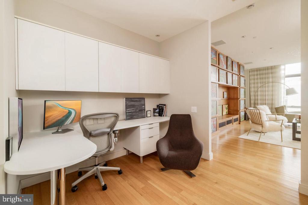 Furnished home office with a curved white desk, built-in cabinets, and hardwood floors adjoining a living area.