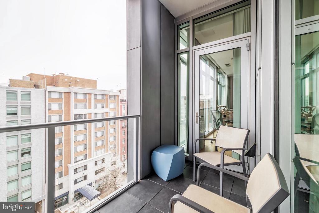 Condo balcony with glass railing, light chairs, and a blue side table overlooking nearby buildings.