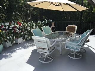 Patio dining set with white wicker chairs beneath a large umbrella, bordered by flowering plants and greenery.