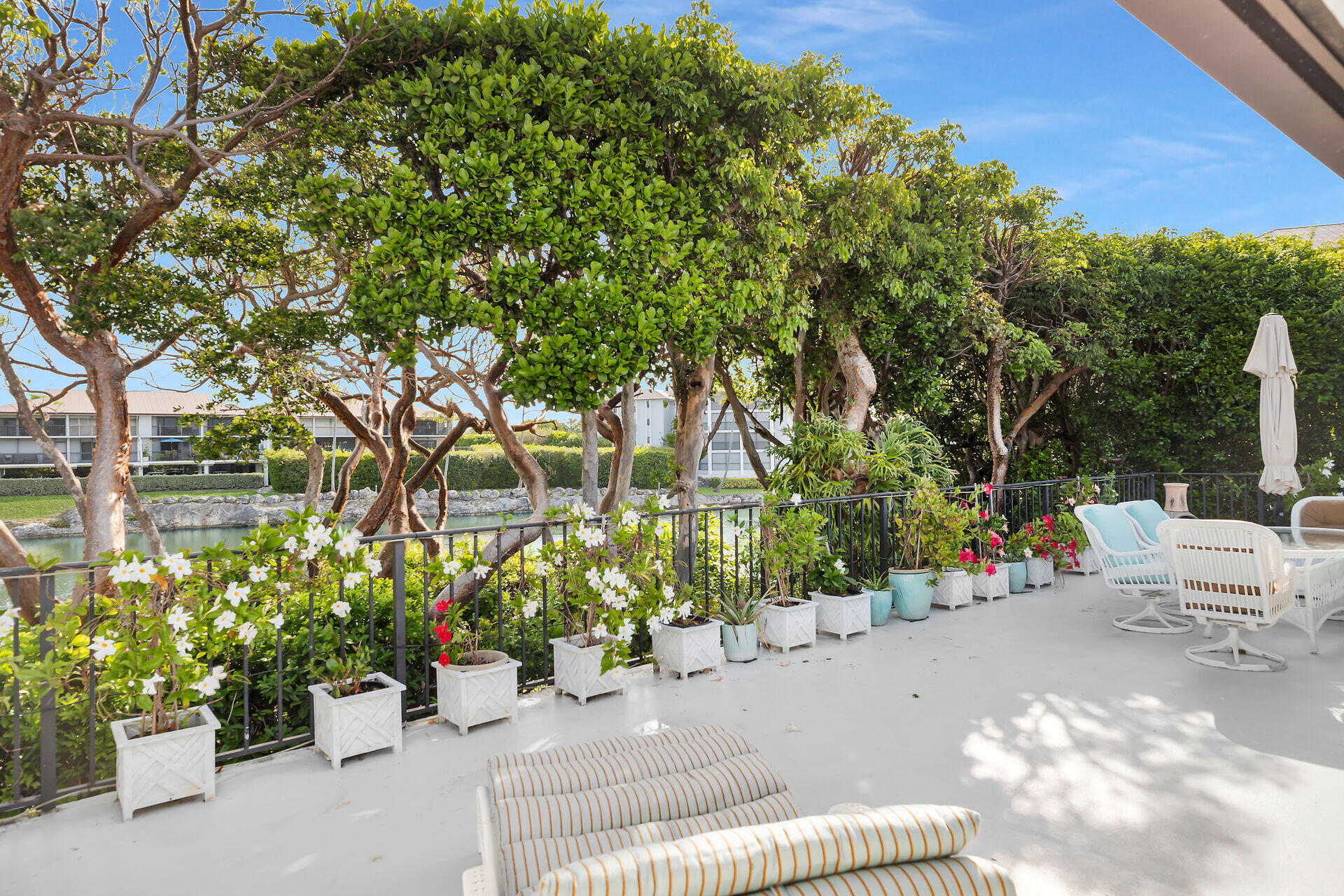 Patio beside a canal, shaded by mature trees and lined with flowering potted plants.