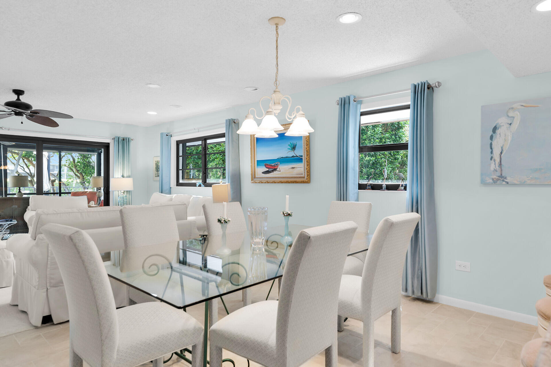 Dining area with a glass-top table, white upholstered chairs, pale blue curtains, and views of greenery.