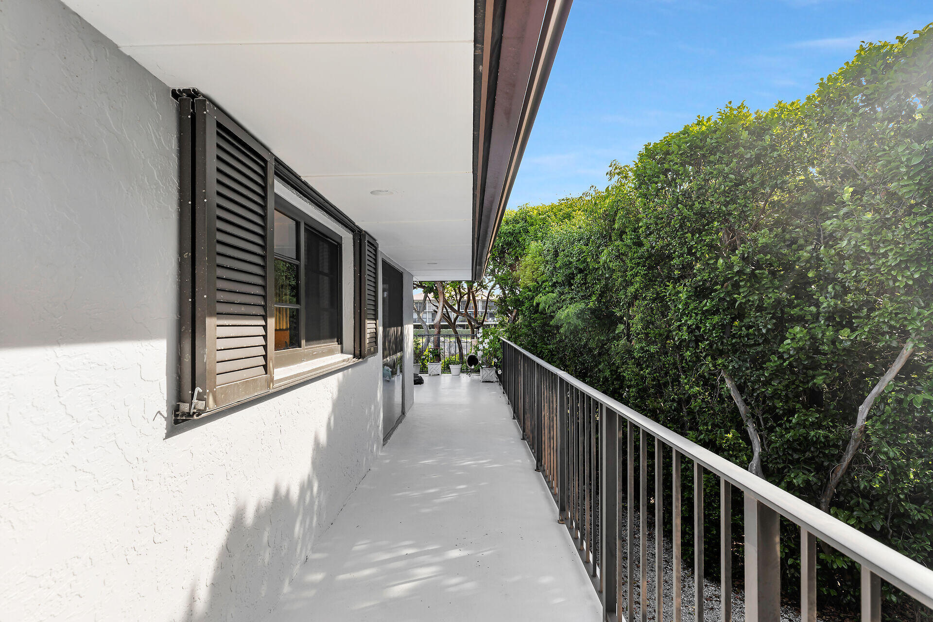 Covered balcony walkway with dark shutters, metal railing, and dense trees alongside the building.