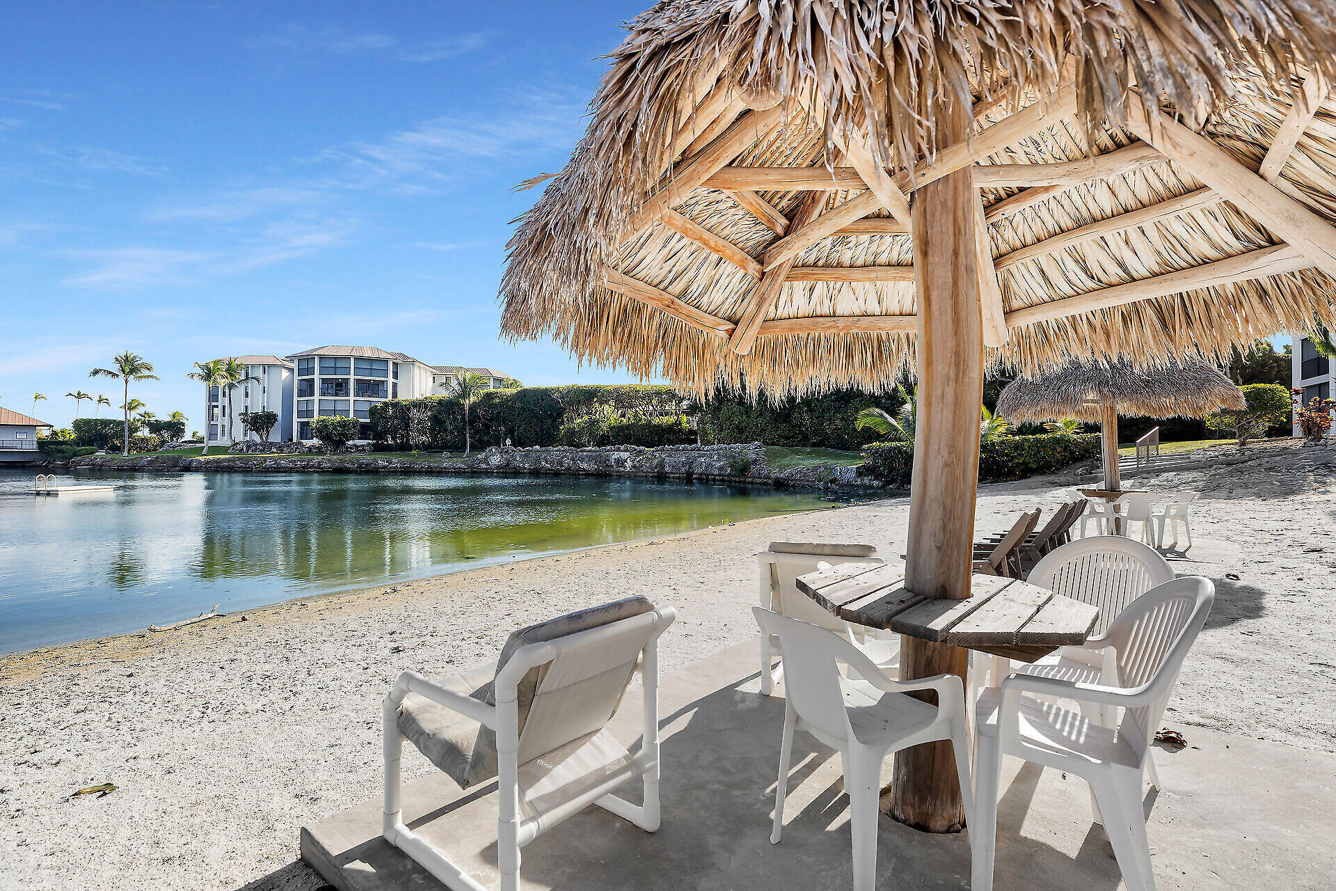 Sandy waterfront beach with a large tiki hut and white seating beside calm water.